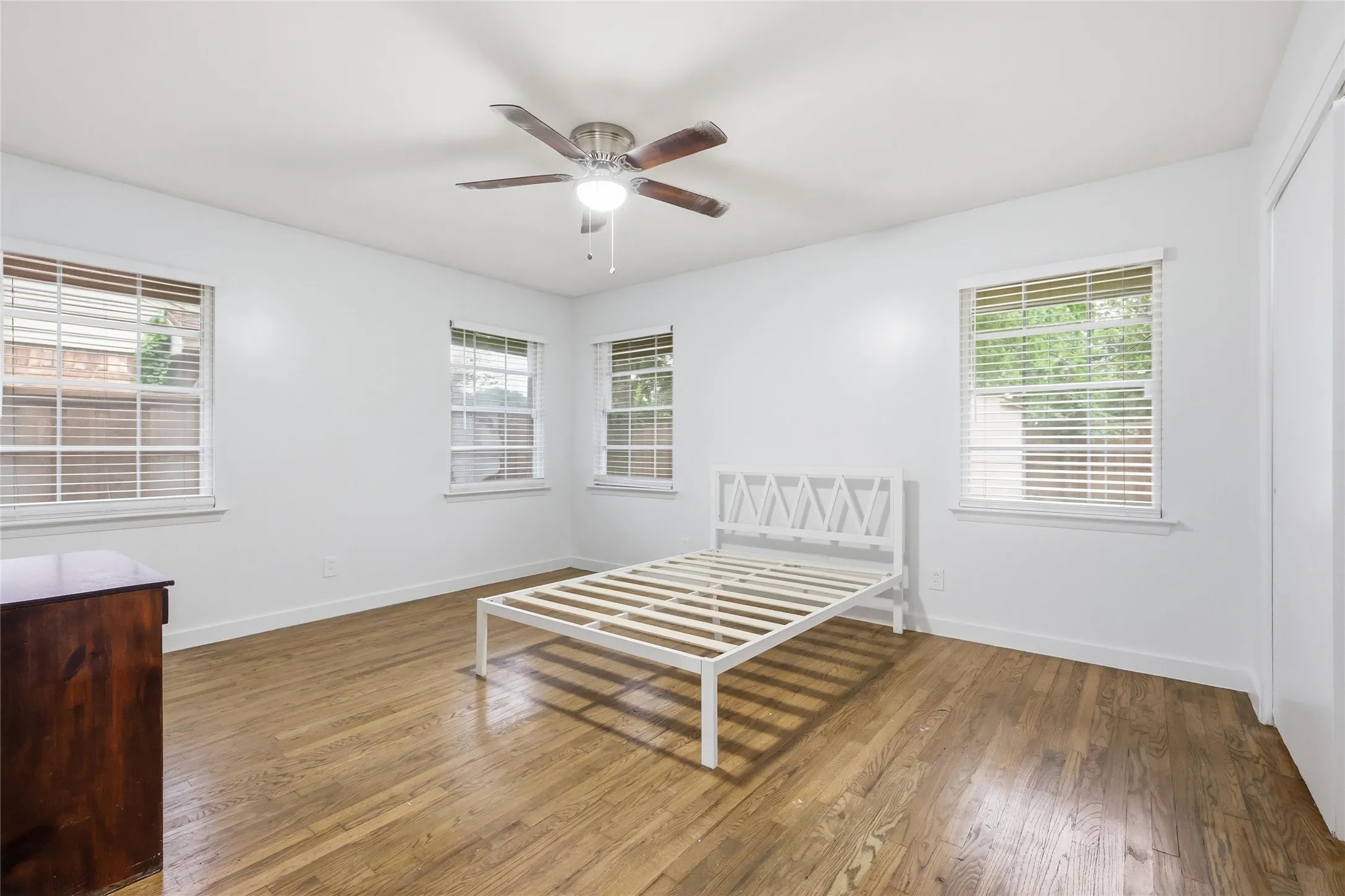 Spacious room featuring wood-finish flooring, multiple windows with blinds, a ceiling fan with integrated lighting, and a white color palette