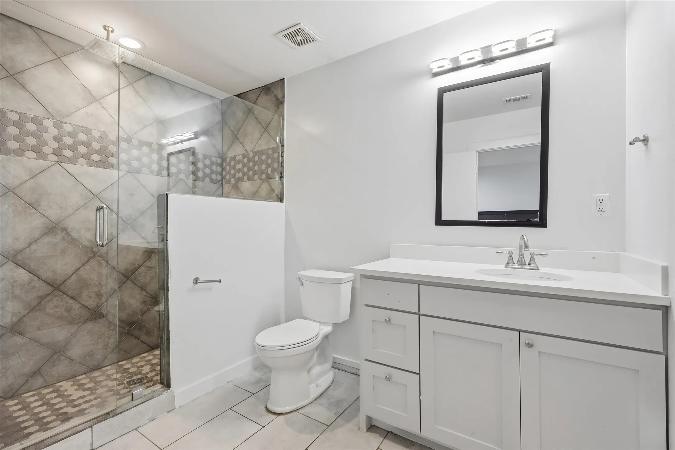 Bathroom featuring a glass-enclosed shower with tiled surround, a white vanity with an integrated sink, a rectangular framed mirror, and tile flooring