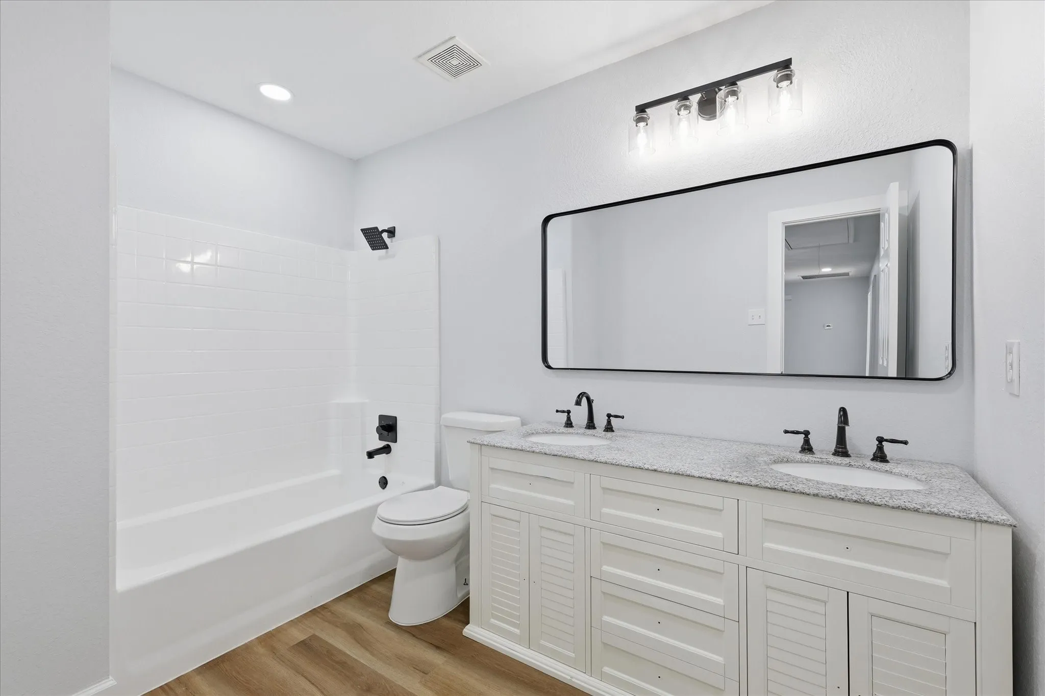 Bathroom featuring a white subway tile tub surround, black hardware, a dual-sink vanity with a speckled countertop, and wood-finish flooring