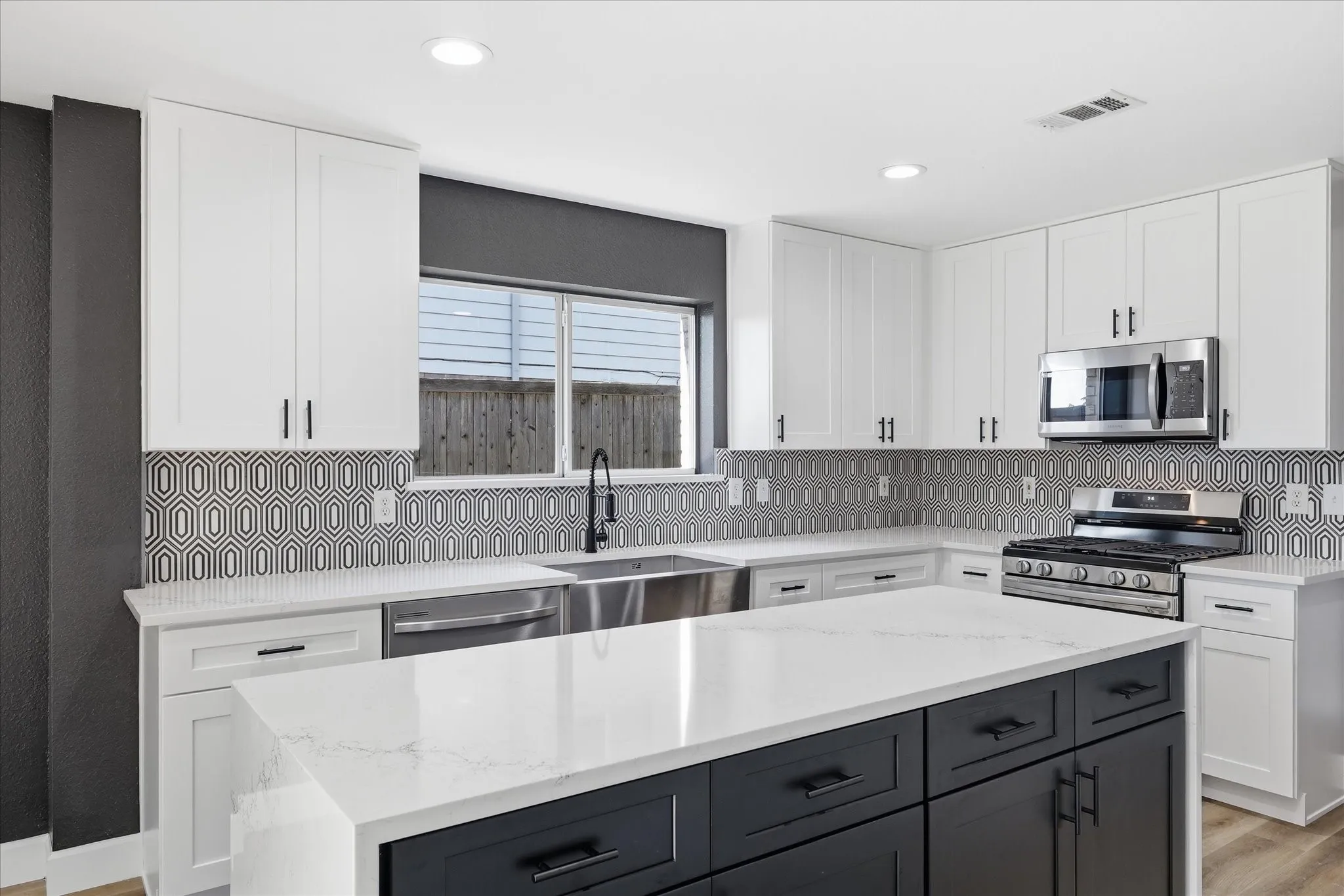 Contemporary kitchen featuring an oversized island with a quartz countertop, sleek white cabinetry, a geometric pattern tile backsplash, a stainless steel gas range, and a built-in microwave