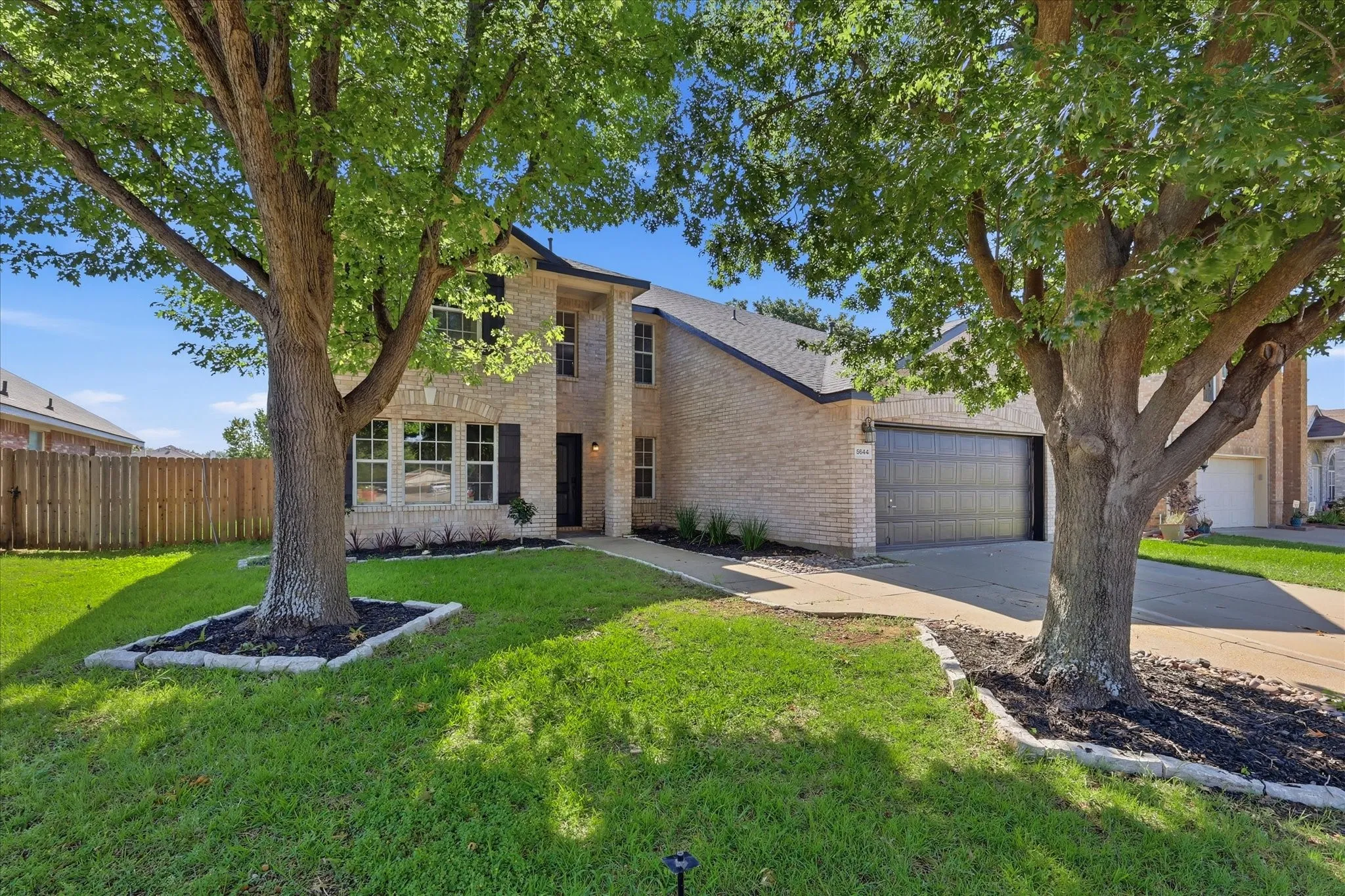 Two-story brick exterior featuring a dark asphalt shingle roof, a two-car garage with a dark panel door, a concrete driveway, and mature trees framing the property