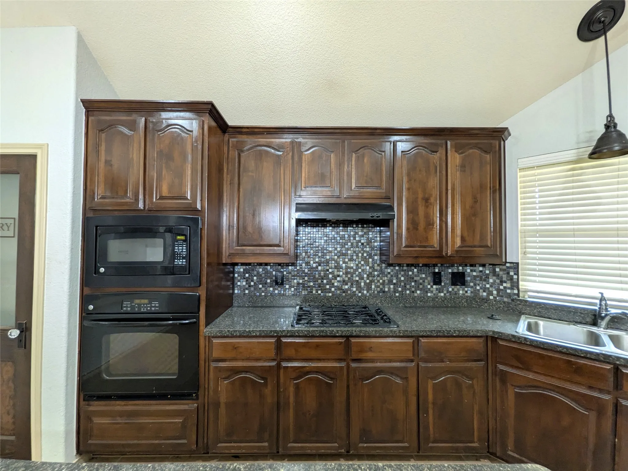 Kitchen featuring dark wood cabinetry, granite-style countertops, mosaic tile backsplash, stainless steel double basin sink, and built-in black appliances