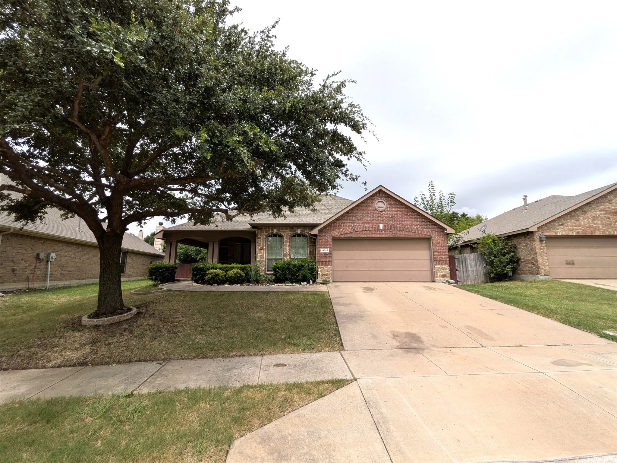 Brick facade home featuring an attached garage, covered front porch, and mature front yard tree