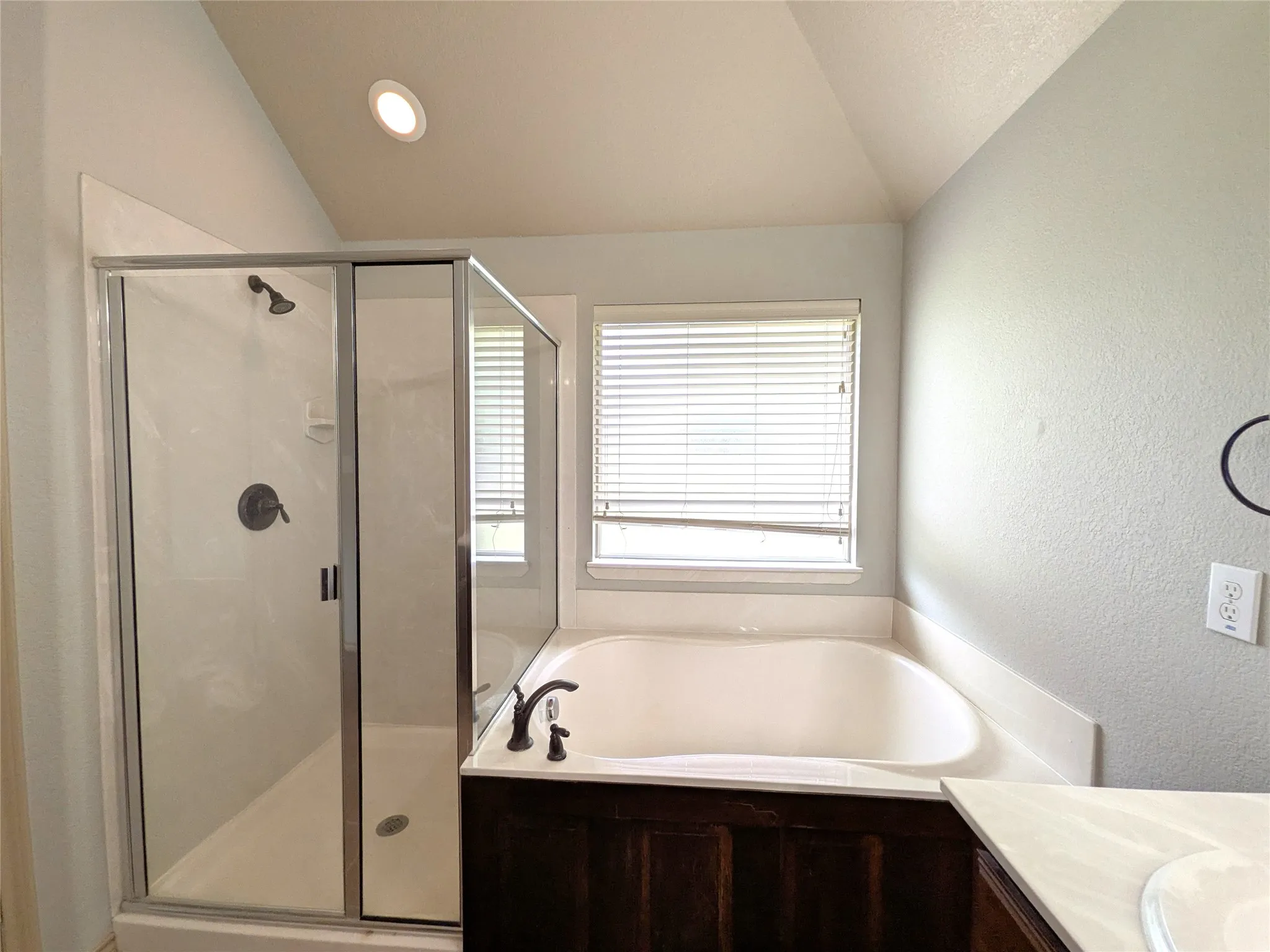 Bathroom featuring a built-in tub, separate glass-enclosed shower with dark bronze fixtures, a window with blinds, and a vanity with dark wood-finish cabinetry and a light countertop