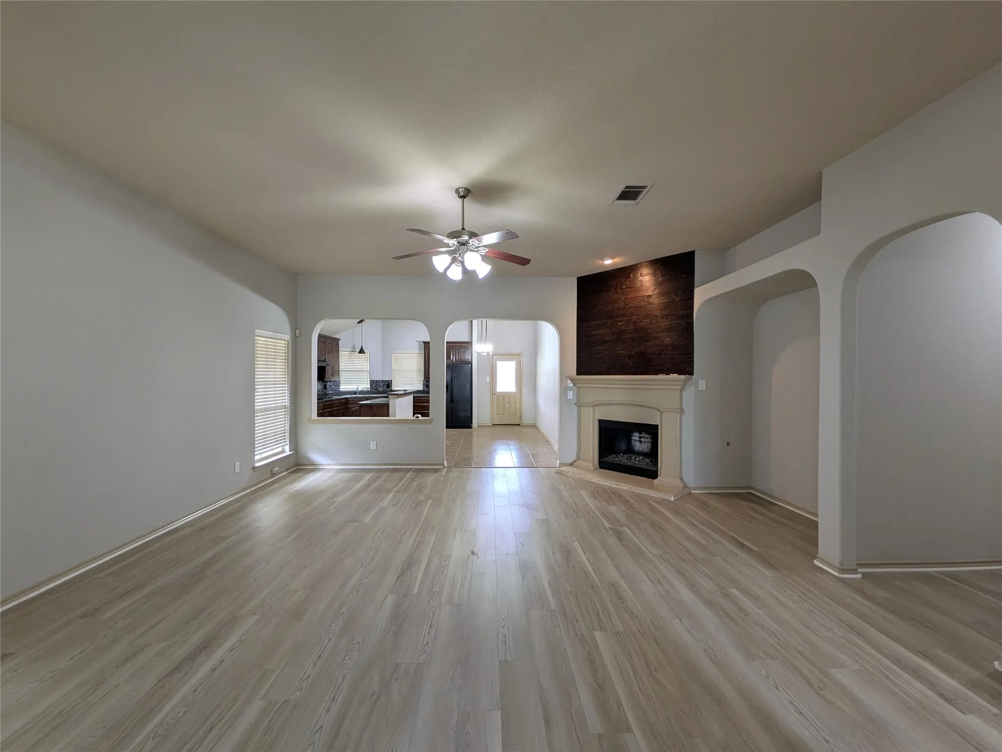 Spacious living area featuring light wood-finish flooring, a contemporary fireplace with a stone-finish mantle, a dark textured accent wall, and arched doorways
