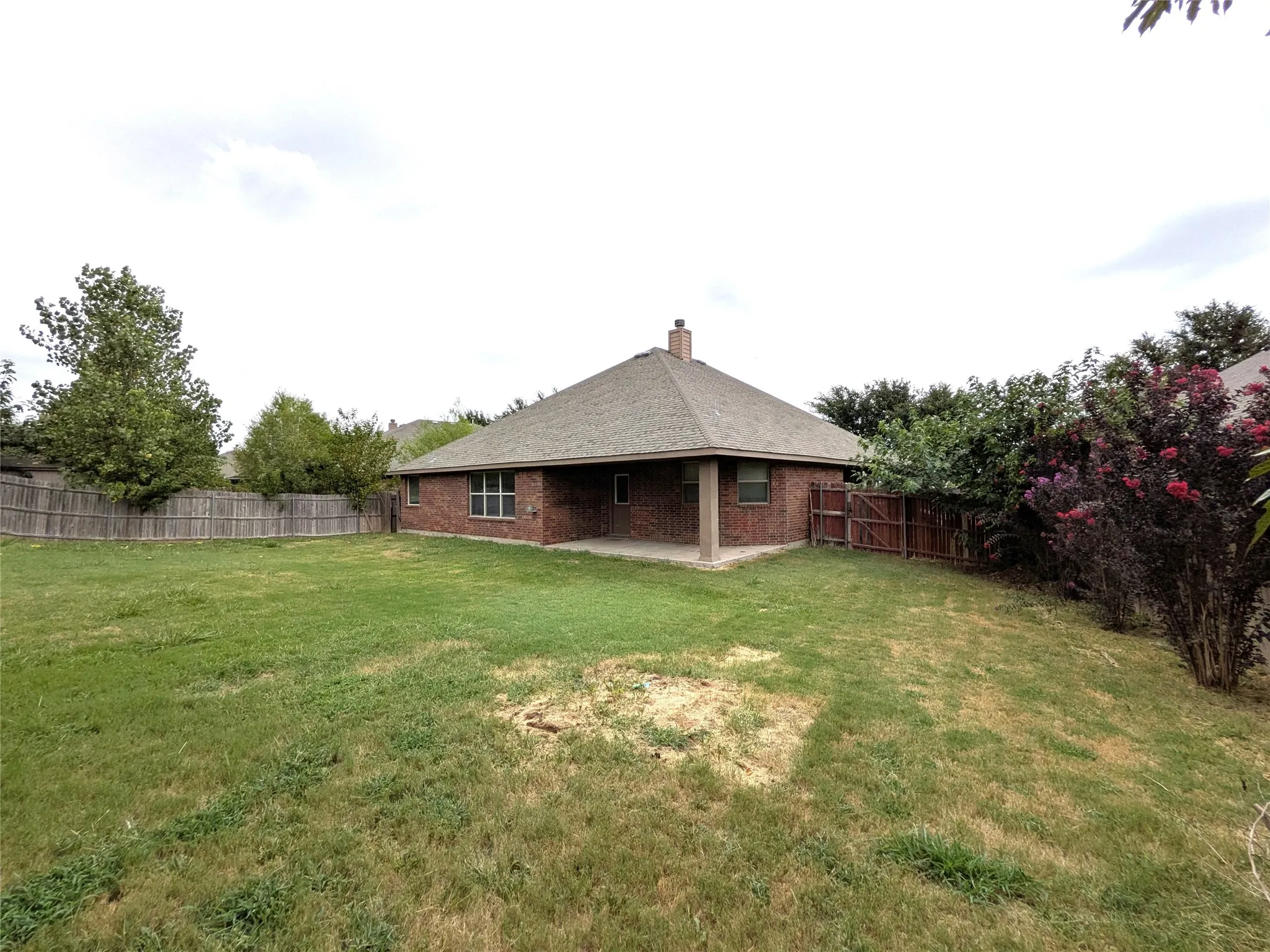Expansive backyard featuring a brick exterior, covered patio, privacy fencing, mature trees, and established shrubs