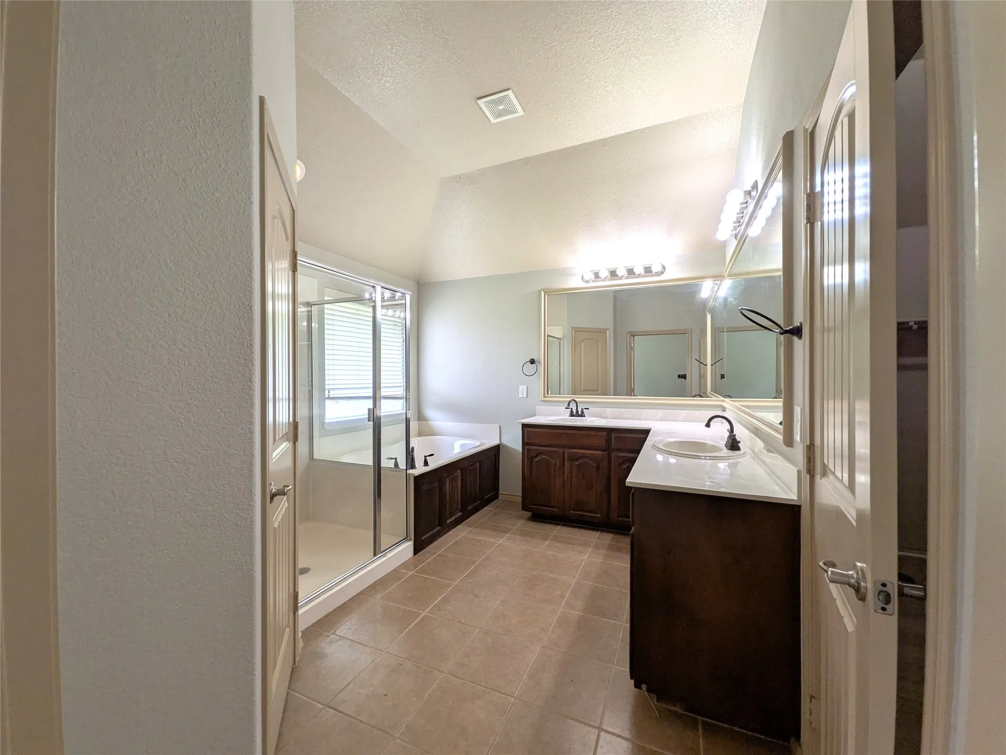 Spacious bathroom featuring dual vanities with white countertops and dark wood cabinetry, a large built-in mirror, a separate glass-enclosed shower, and a built-in bathtub