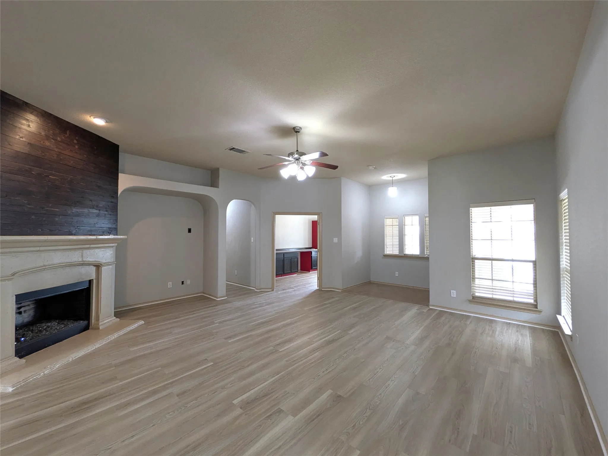 Spacious room featuring light wood-finish flooring, a fireplace with a light-colored stone mantel and hearth, and a dark wood-paneled accent wall