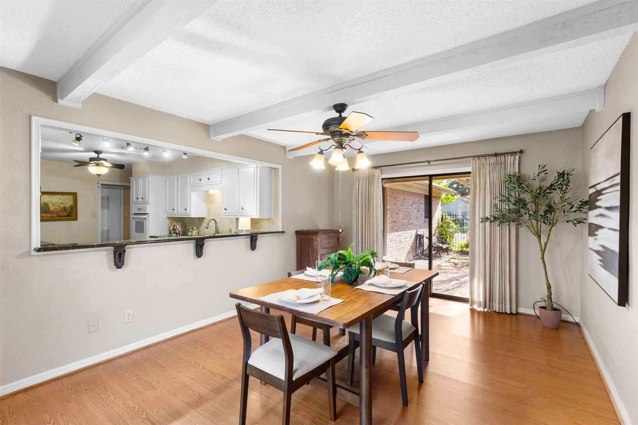Wood-finish flooring, white exposed ceiling beams, recessed kitchen pass-through with built-in bar seating, ceiling fan with light fixture, and sliding glass door