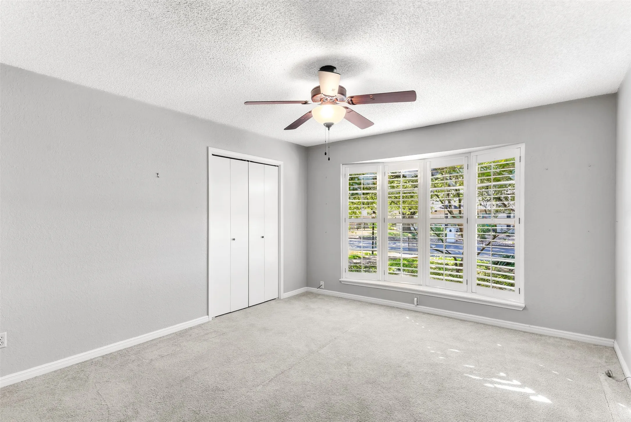 Neutral wall color, ceiling fan with integrated lighting, carpet flooring, bi-fold closet doors, and a multi-pane window with plantation shutters