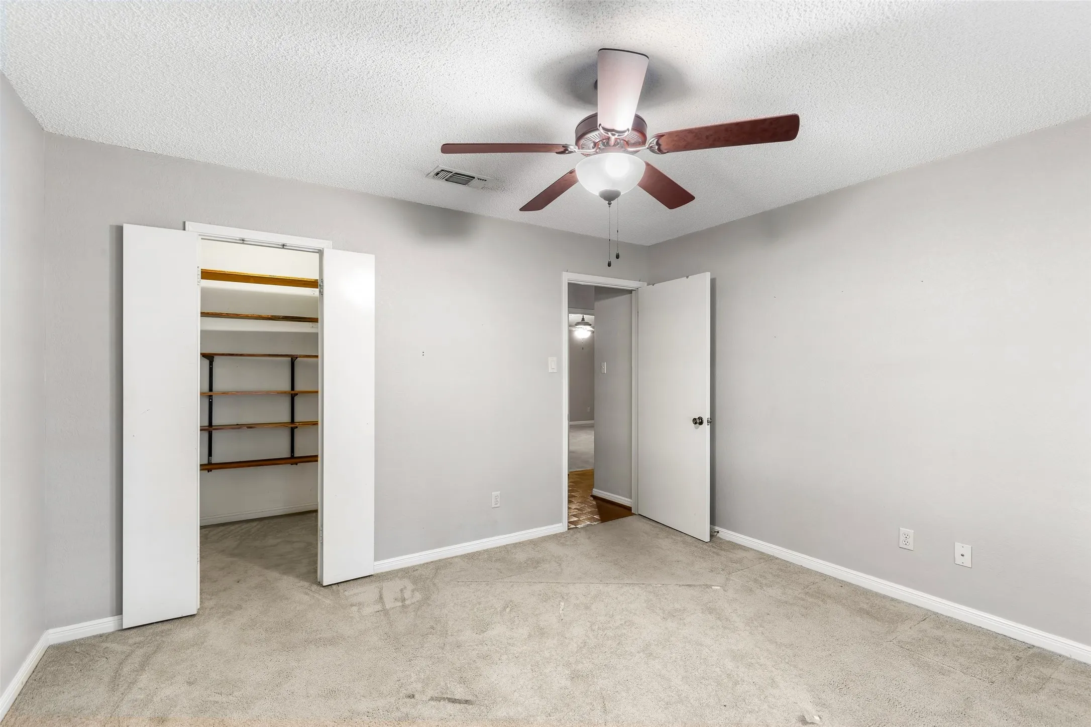 Neutral gray wall paint, textured ceiling, a ceiling fan with light fixture, light gray carpet flooring, and a closet with bifold doors and built-in shelving