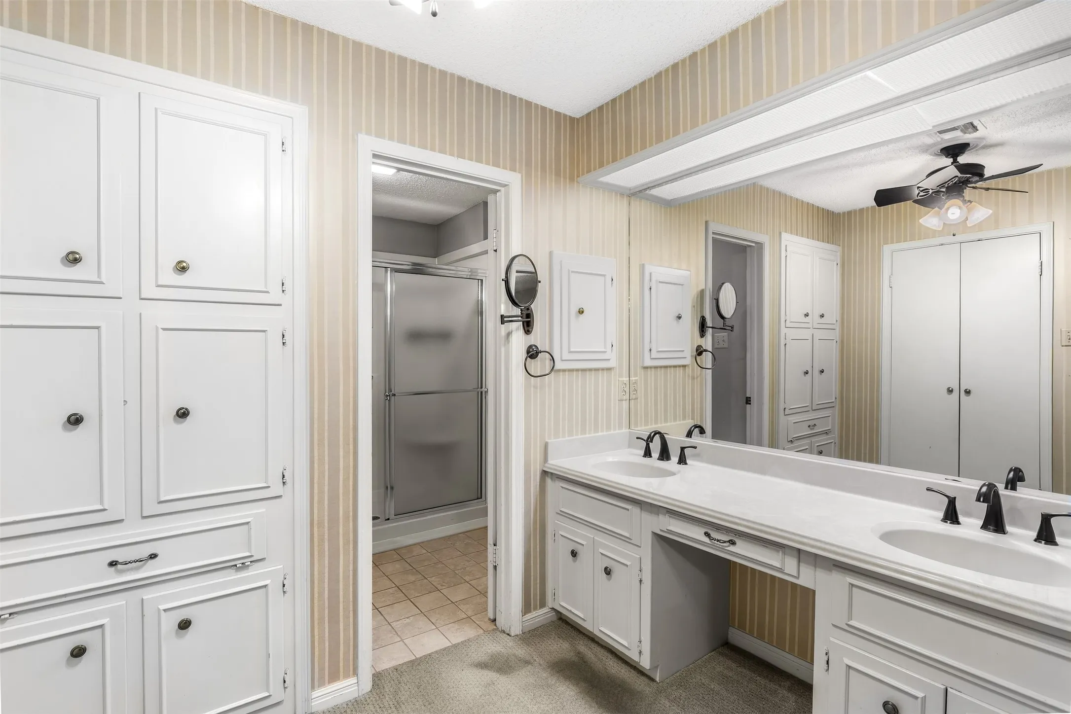 Dual vanity with integrated sinks, built-in cabinetry, full-width mirrored wall, ceiling fan, and walk-in shower with frosted glass enclosure