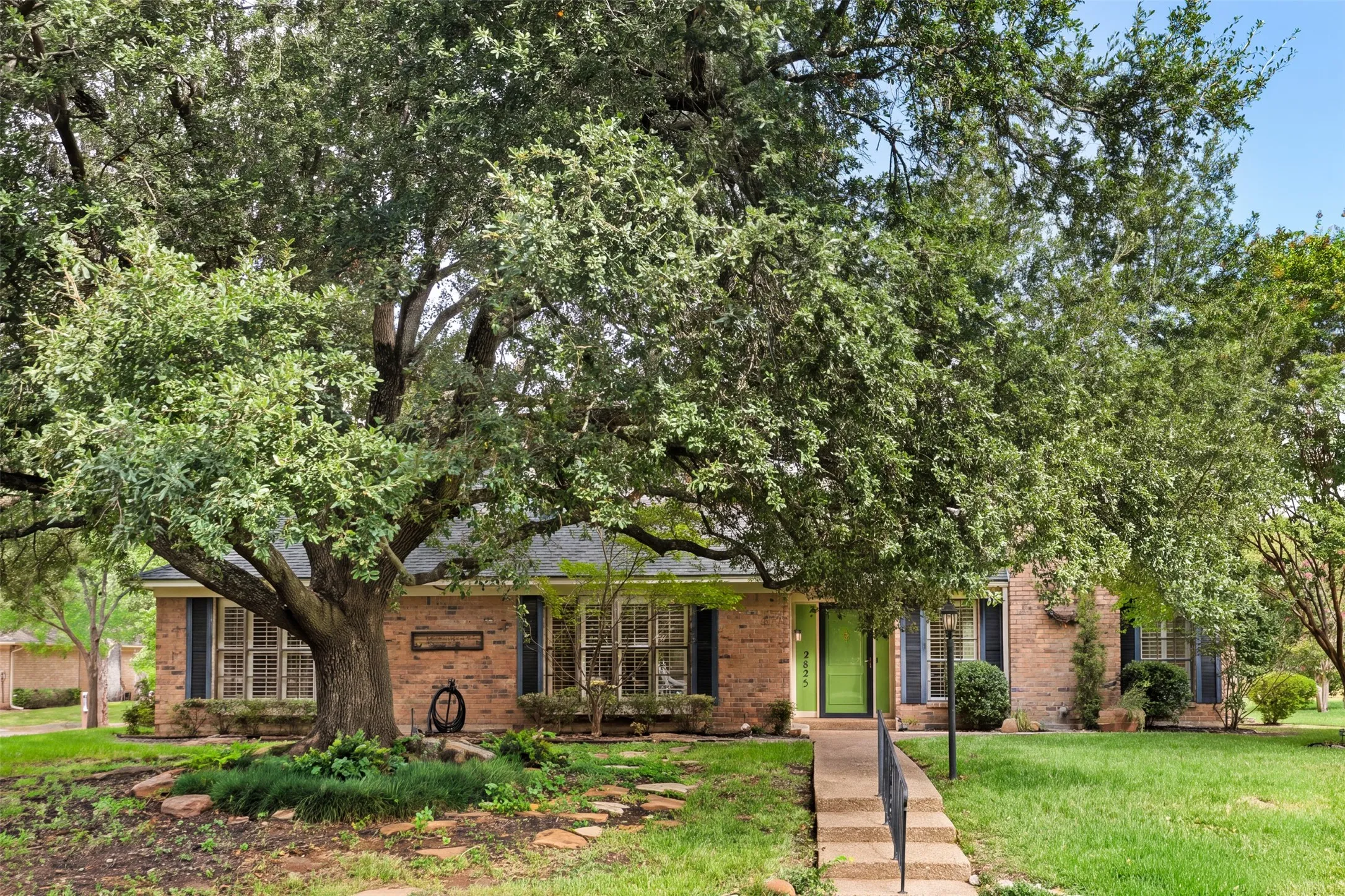 Brick facade residence with mature trees, a stone pathway, a bright green entry door, and black louvered shutters