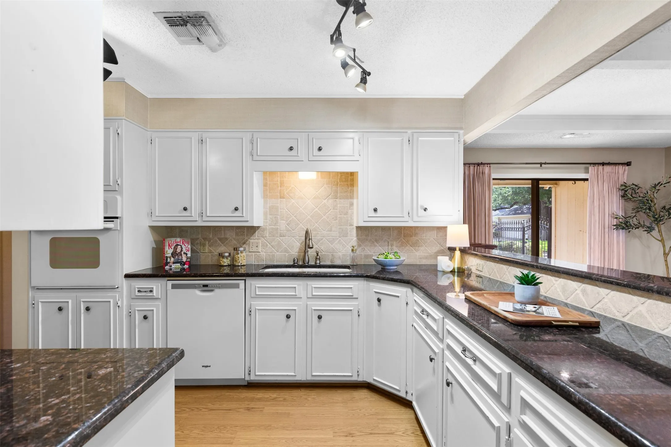 White cabinetry with dark hardware, dark countertops, built-in dishwasher, tile backsplash, and light wood-finish flooring