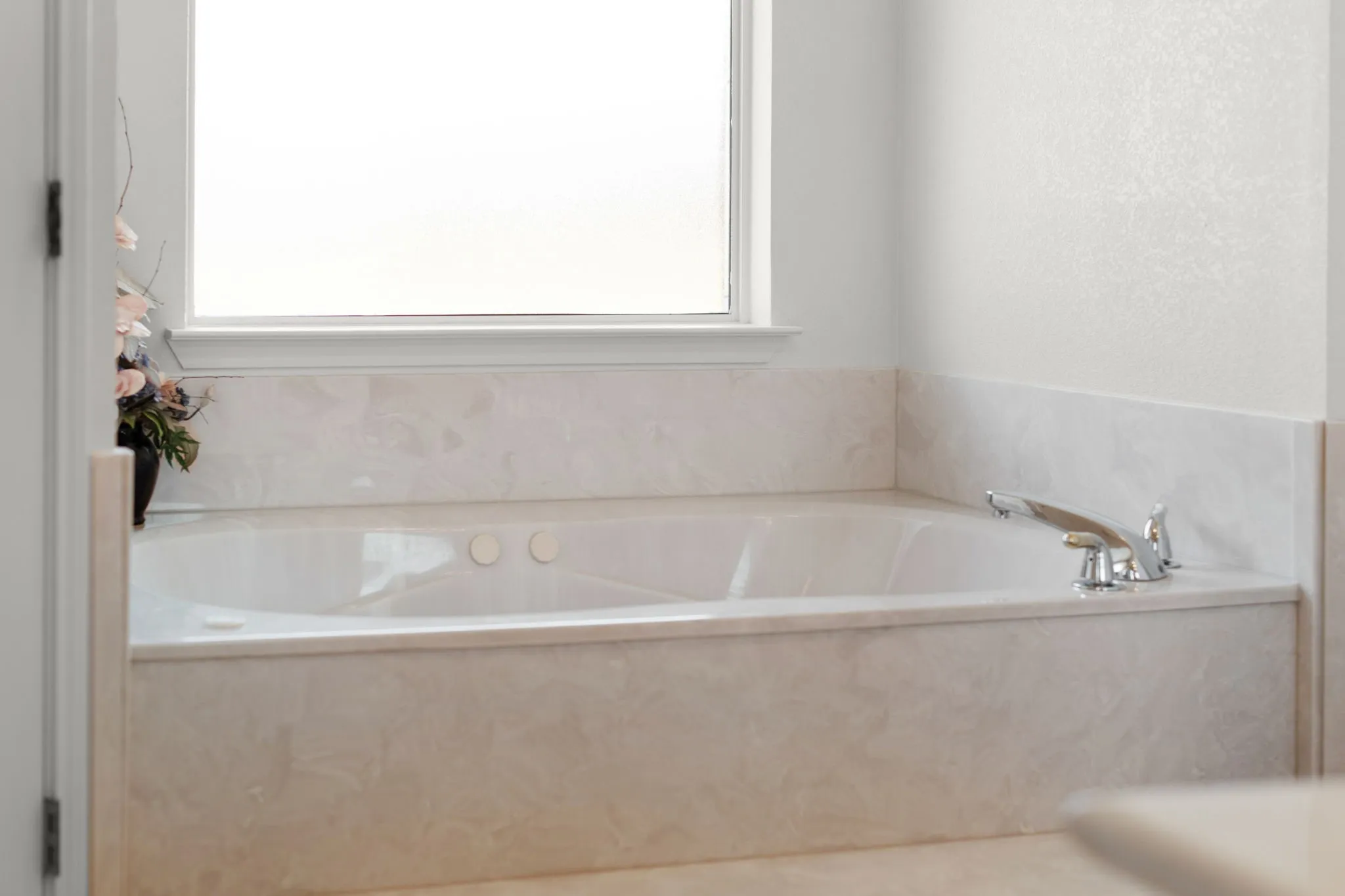 White soaking tub with integrated jets, surrounded by a light-toned stone-finish deck and apron, beneath a frosted window