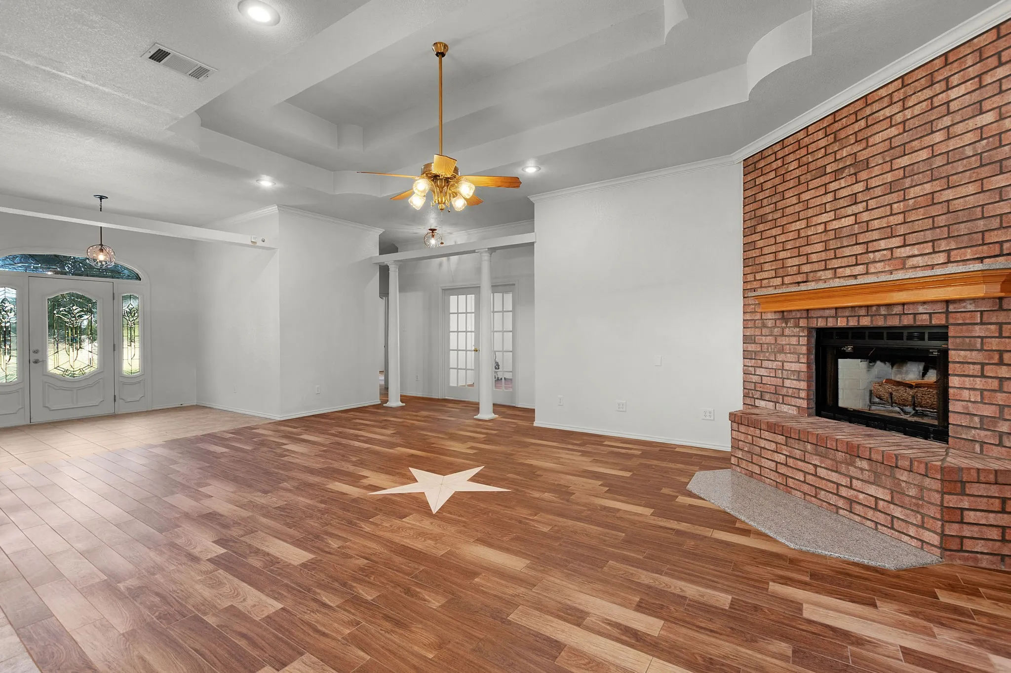 Spacious room featuring wood-finish flooring with an inlaid star design, a prominent brick fireplace with a wood mantel, a tray ceiling with recessed lighting and a ceiling fan, and double French doors with decorative glass