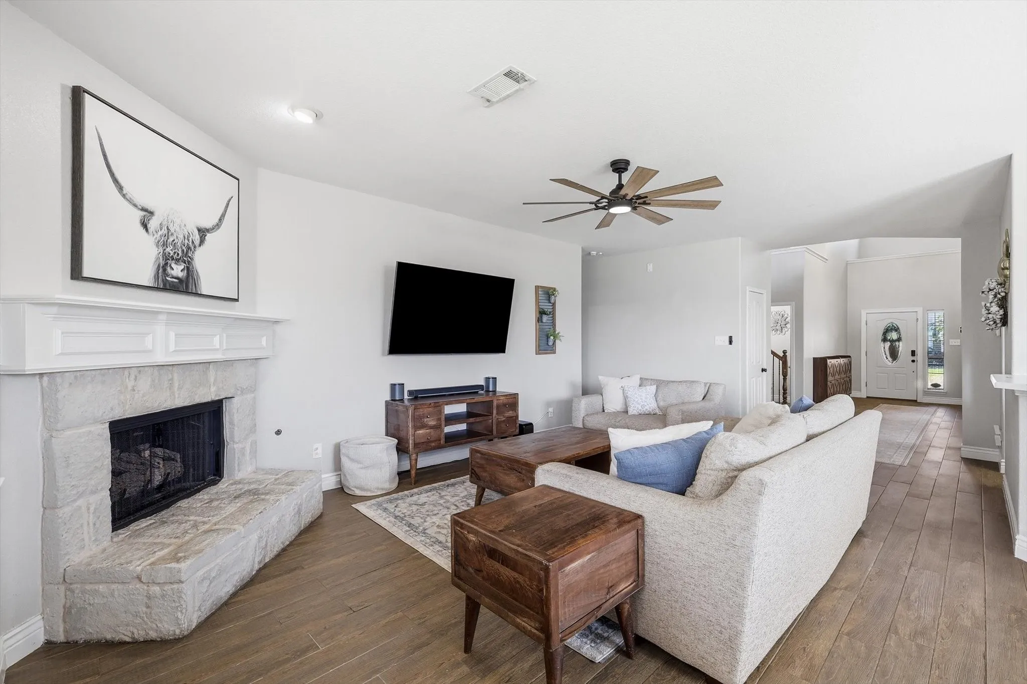 Spacious living area featuring a stone fireplace with a white mantel, wood-finish flooring, and a ceiling fan