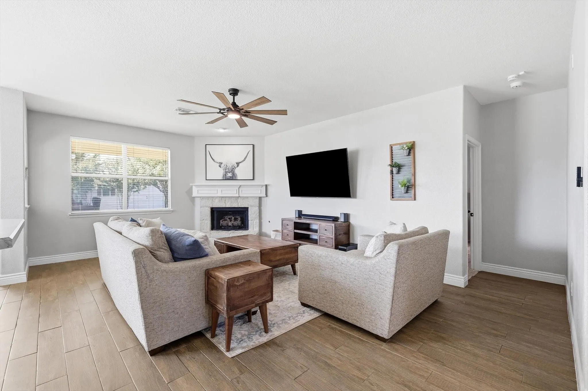 Spacious living area featuring light wood-finish flooring, a white mantel fireplace, and a ceiling fan with integrated lighting