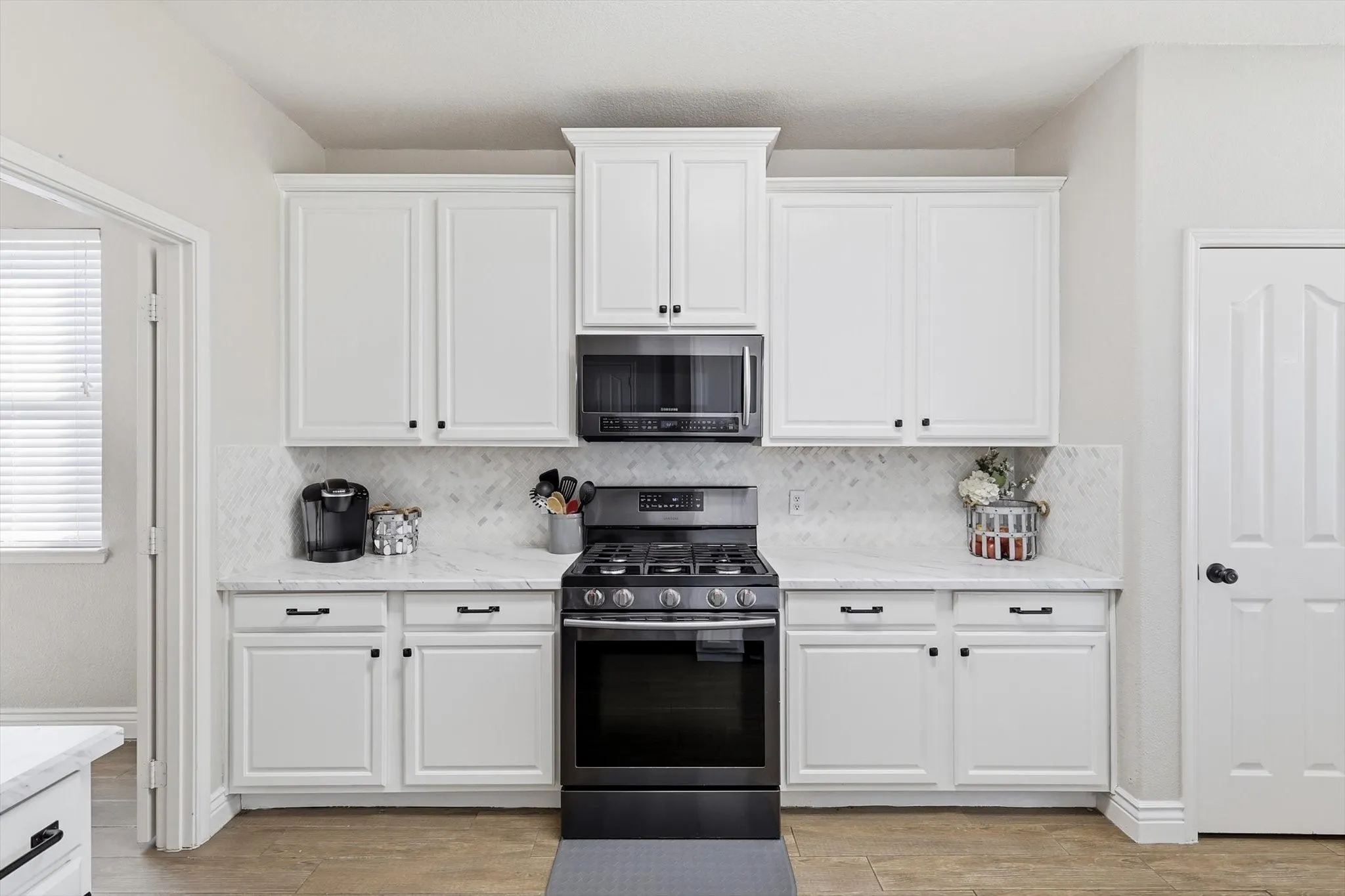 Kitchen featuring white shaker-style cabinetry with matte black hardware, a black gas range with a matching over-the-range microwave, light-toned countertops, and a herringbone pattern tile backsplash