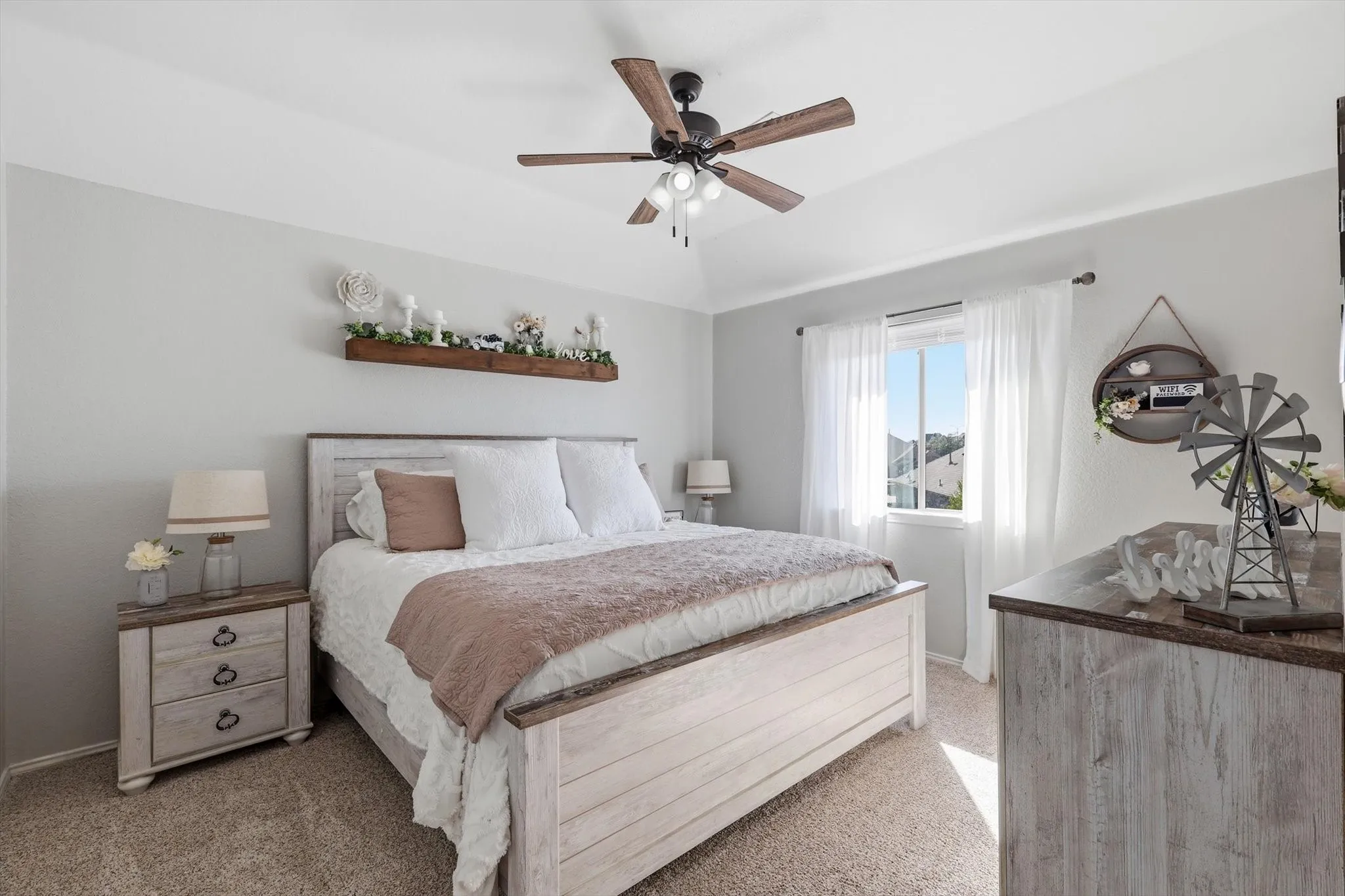 Carpeted room featuring a ceiling fan with light fixture, a window with white curtains, a built-in wall shelf, and light gray wall paint