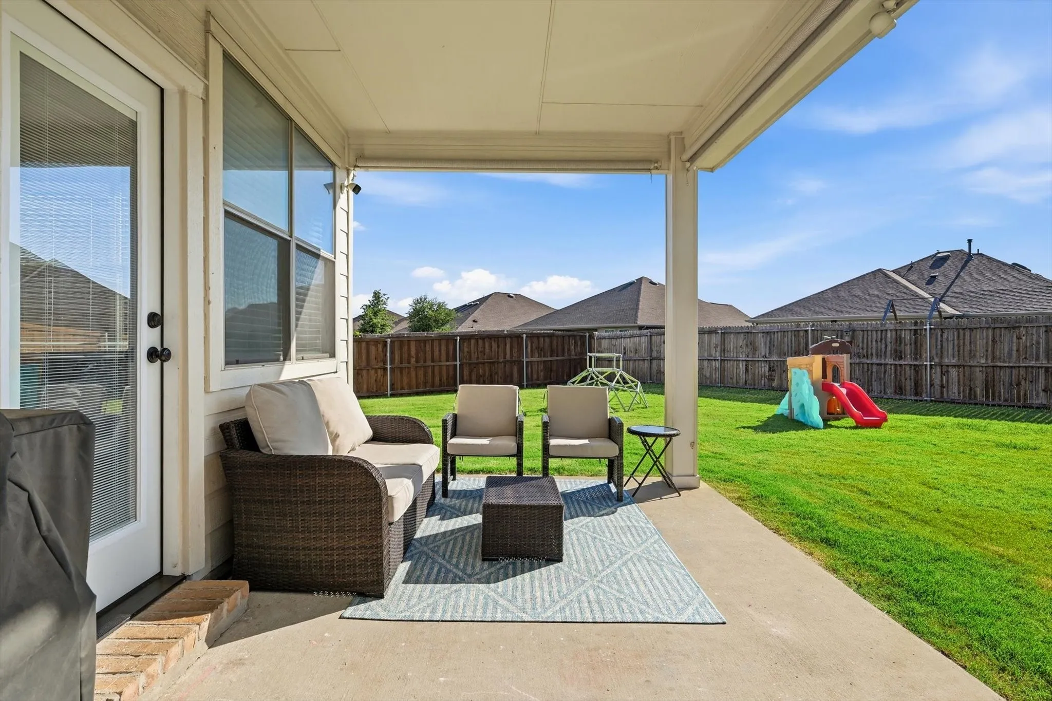 Expansive concrete patio beneath a covered extension, featuring a white door with glass panels, an adjacent window with blinds, and direct access to a fenced backyard with a manicured lawn