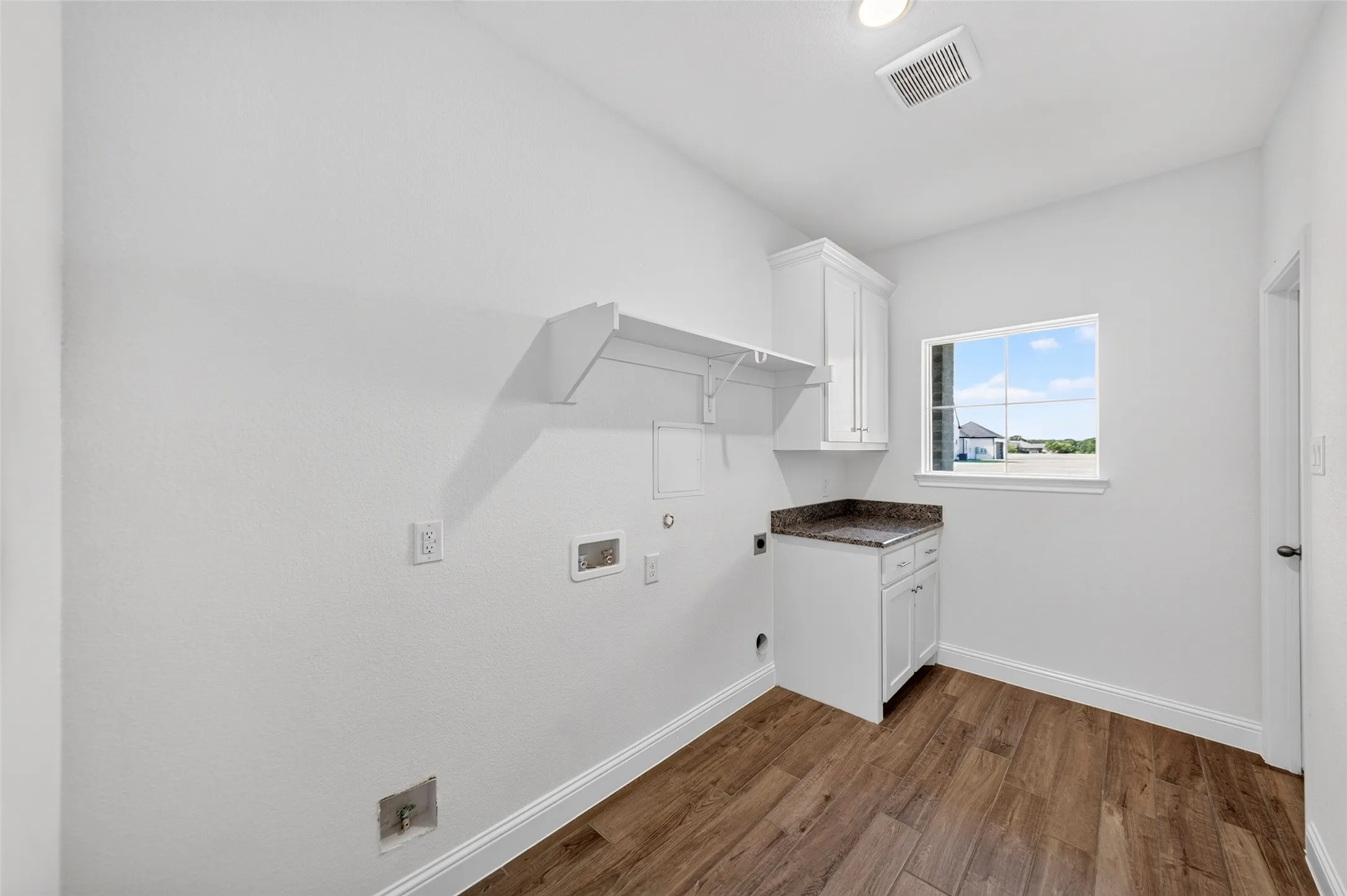 Dedicated utility space featuring wood-finish flooring, a single window, white base cabinetry with a stone-finish countertop, and wall-mounted shelving