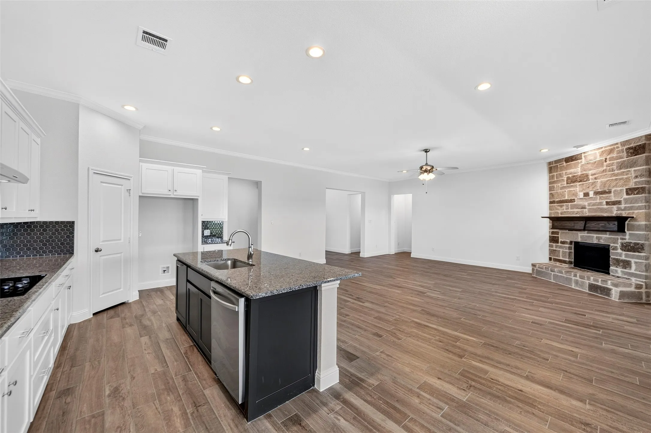 Open-concept interior featuring wood-finish flooring, a kitchen island with sink and stainless steel dishwasher, white cabinetry, recessed lighting, and a stone-clad fireplace