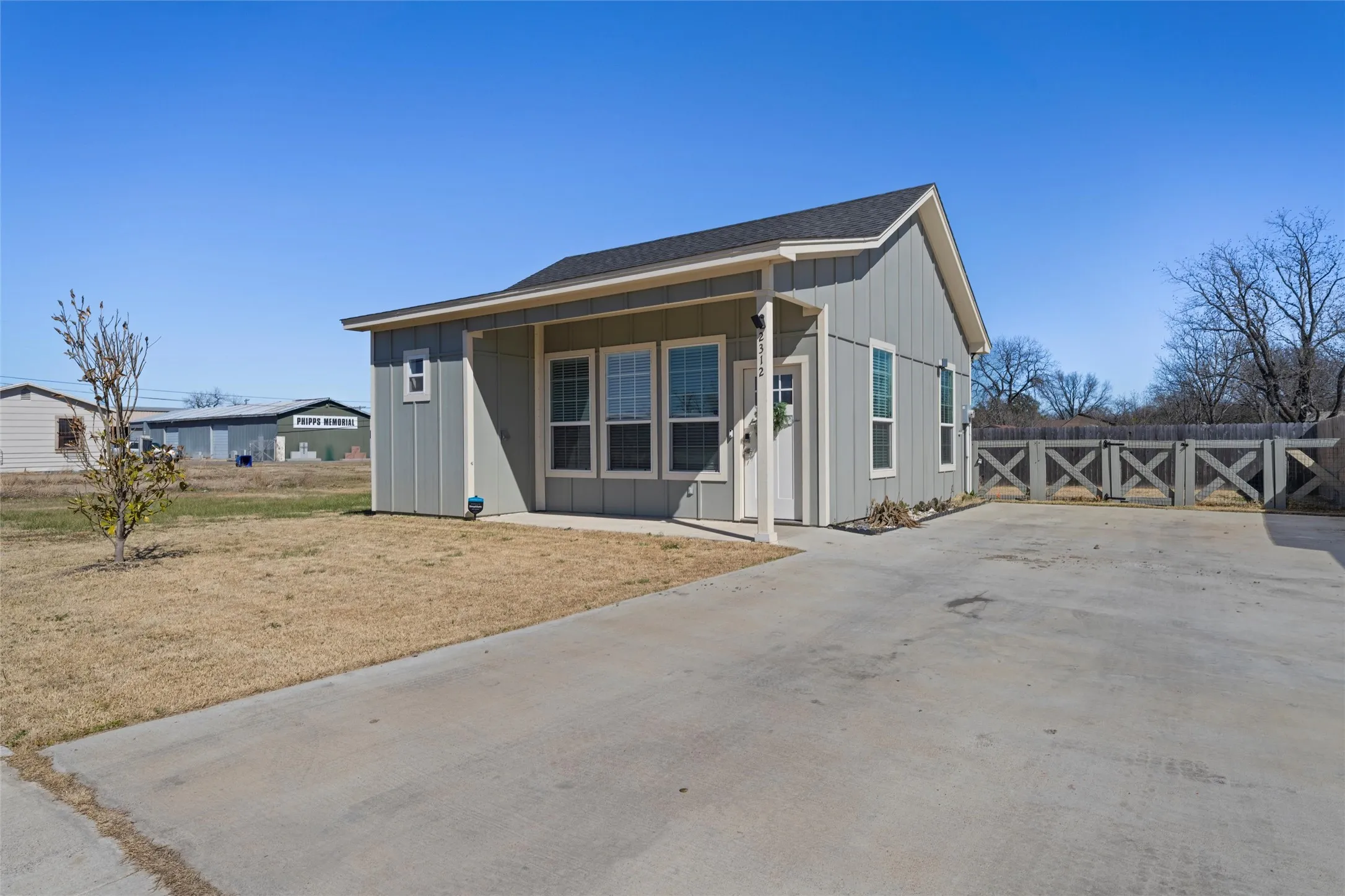 Exterior featuring vertical siding in a neutral tone, an asphalt shingle roof, a covered entry porch, a concrete driveway, and a decorative wood fence along the property line