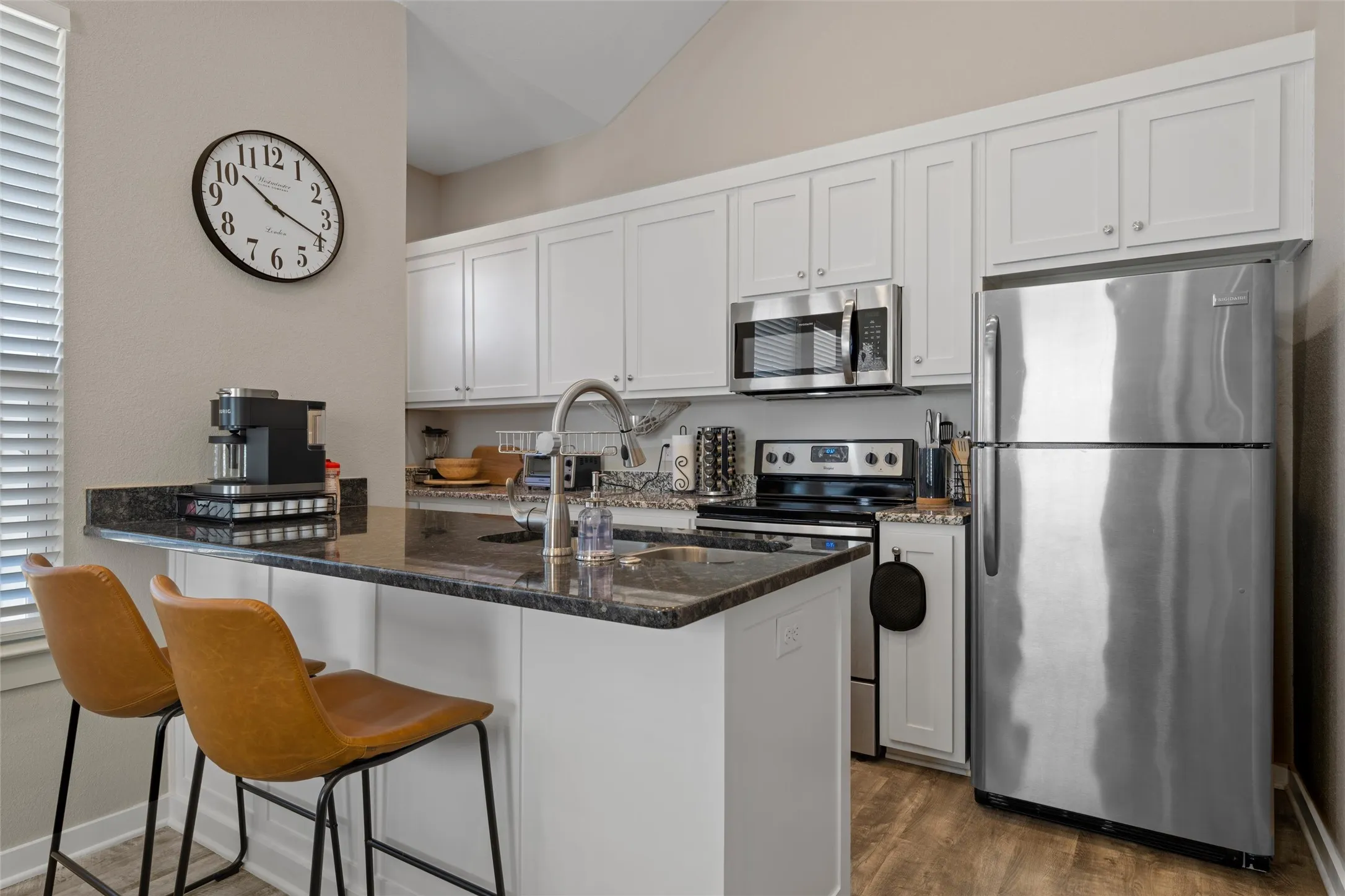 Modern kitchen featuring granite countertops, white cabinetry, stainless steel appliances, wood-finish flooring, and a breakfast bar