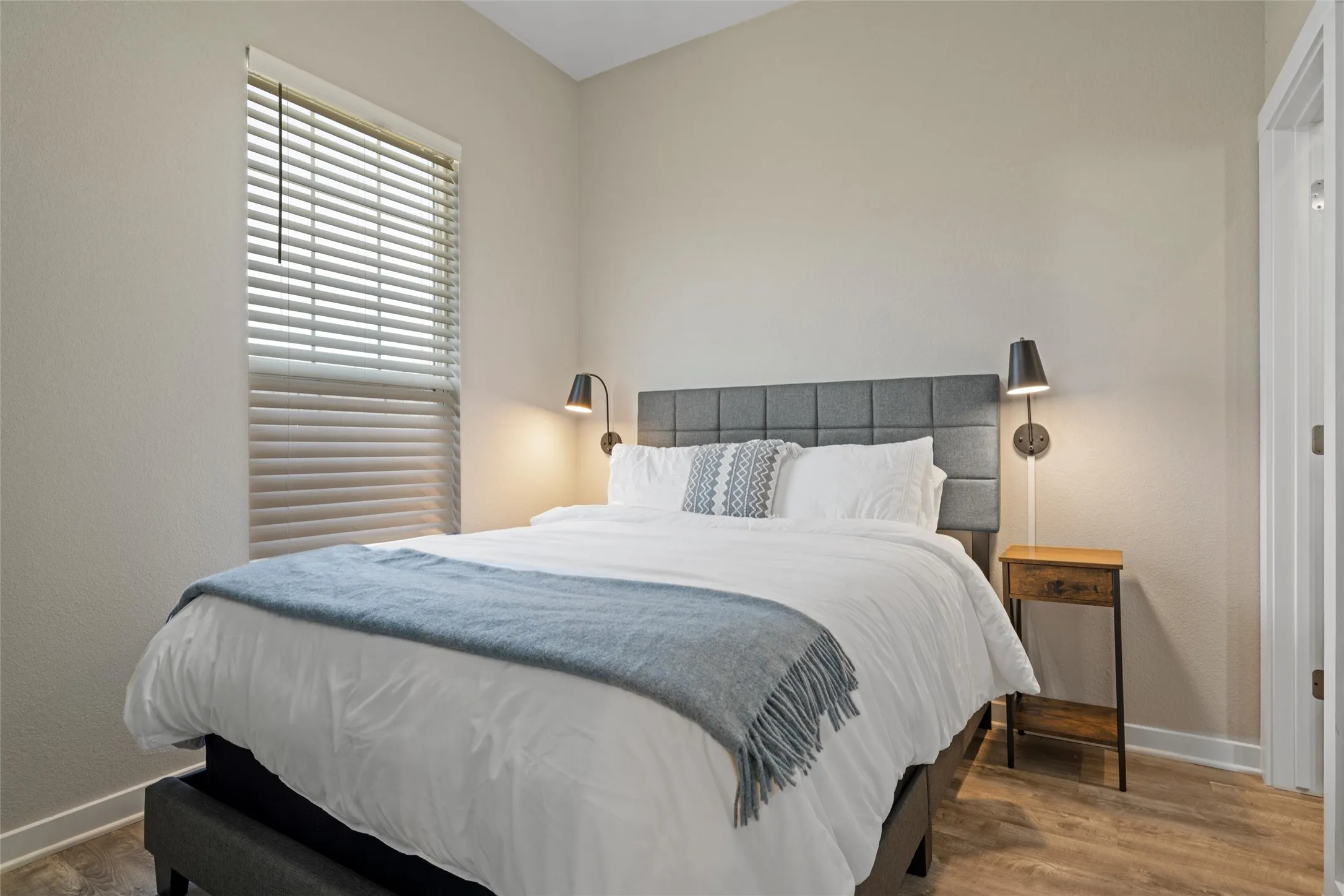 Bedroom featuring wood-finish flooring, a window with horizontal blinds, a paneled headboard, and wall-mounted sconces