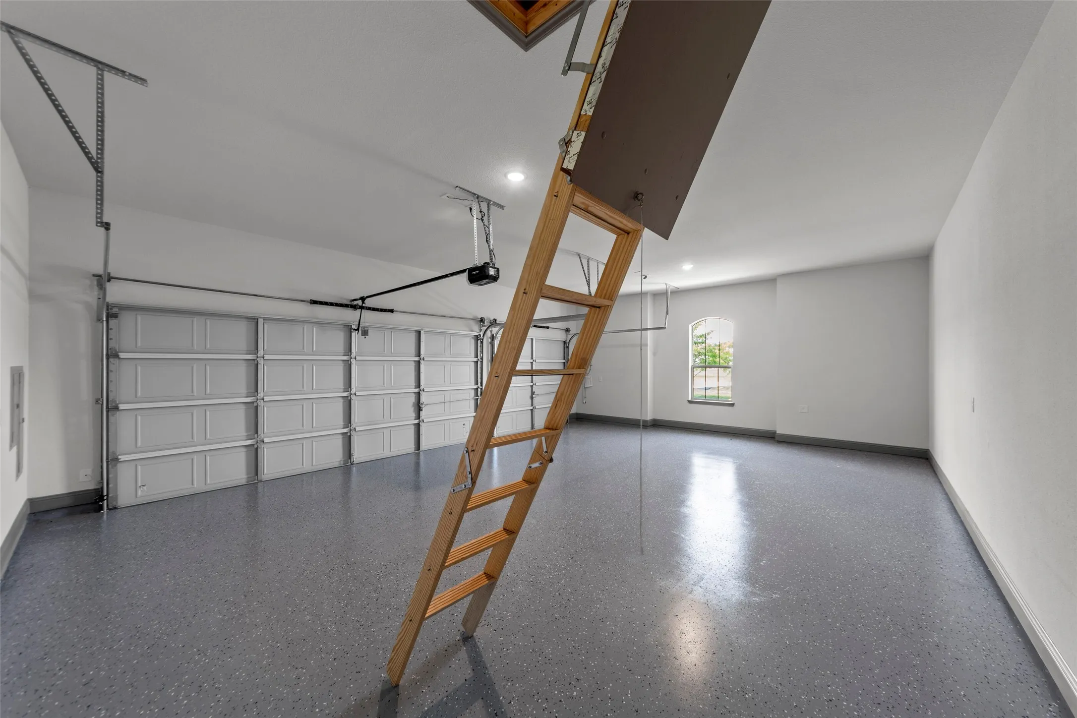Spacious garage featuring a durable epoxy-coated floor, automatic garage door opener, attic access ladder, recessed lighting, and an arched window