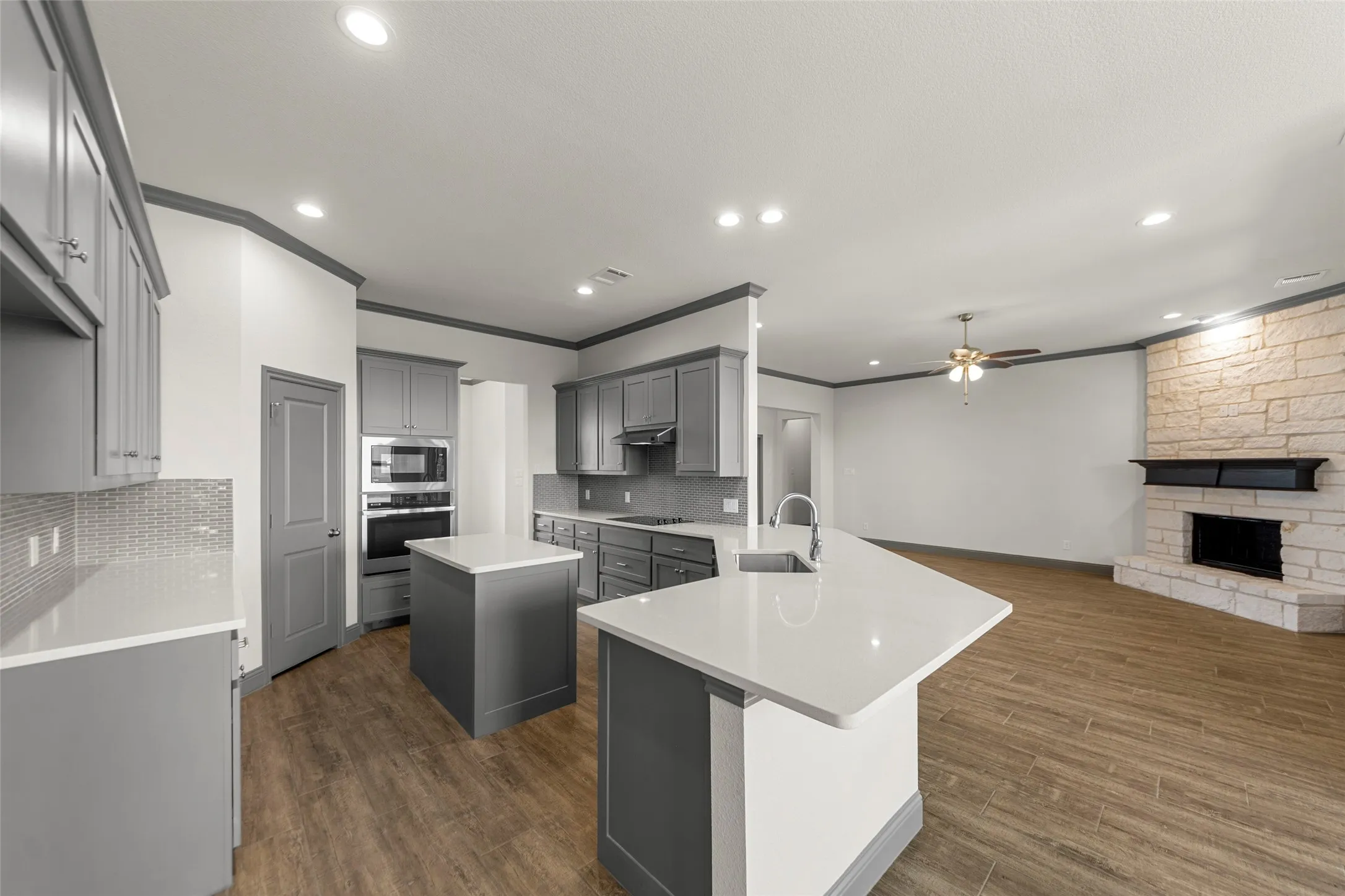 Open-concept kitchen and living area featuring wood-finish flooring, recessed lighting, and a stone-clad fireplace with a dark wood mantle