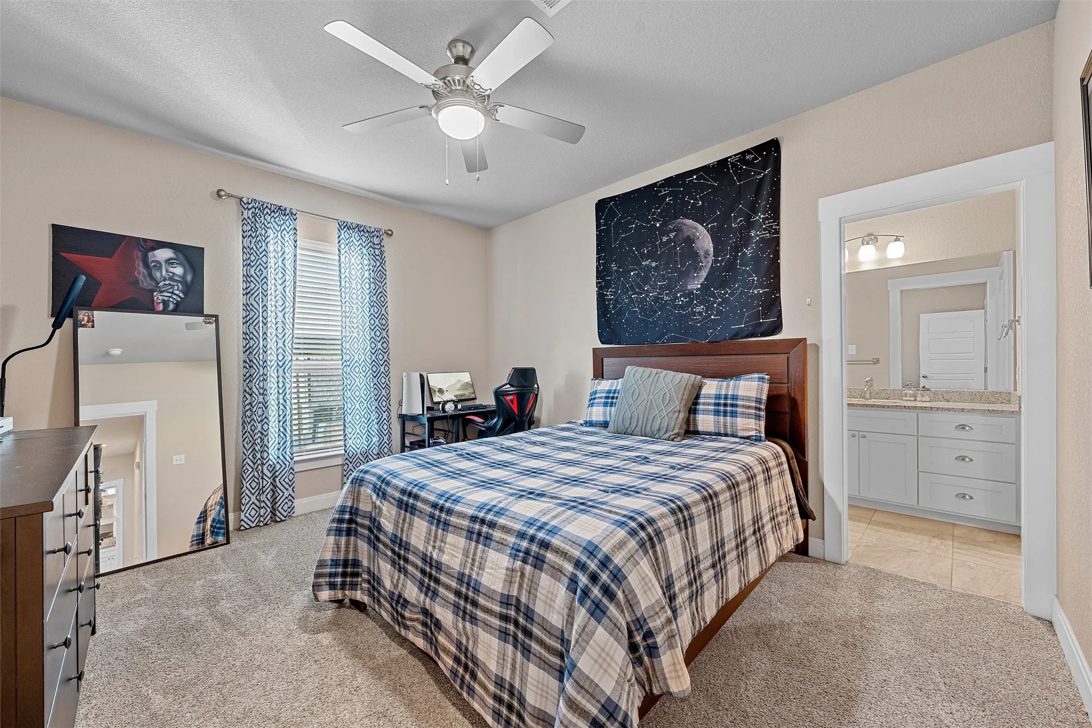 Carpeted bedroom featuring a ceiling fan with integrated lighting, a window with blinds, and an open doorway revealing a bathroom with a vanity and tile flooring