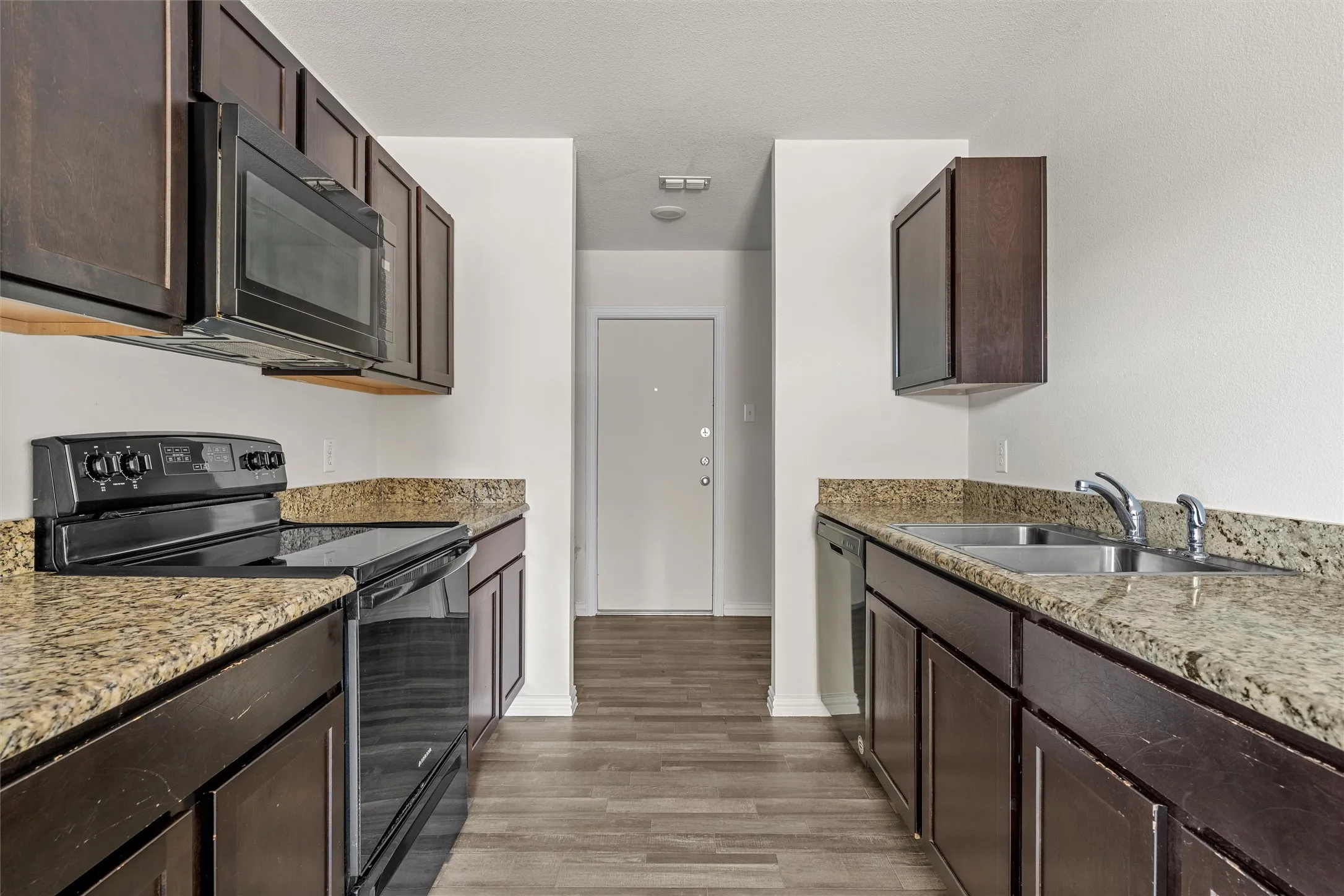Galley kitchen featuring dark wood-finish cabinetry, light-toned countertops, wood-finish flooring, a stainless steel double basin sink, and black appliances including a range and microwave