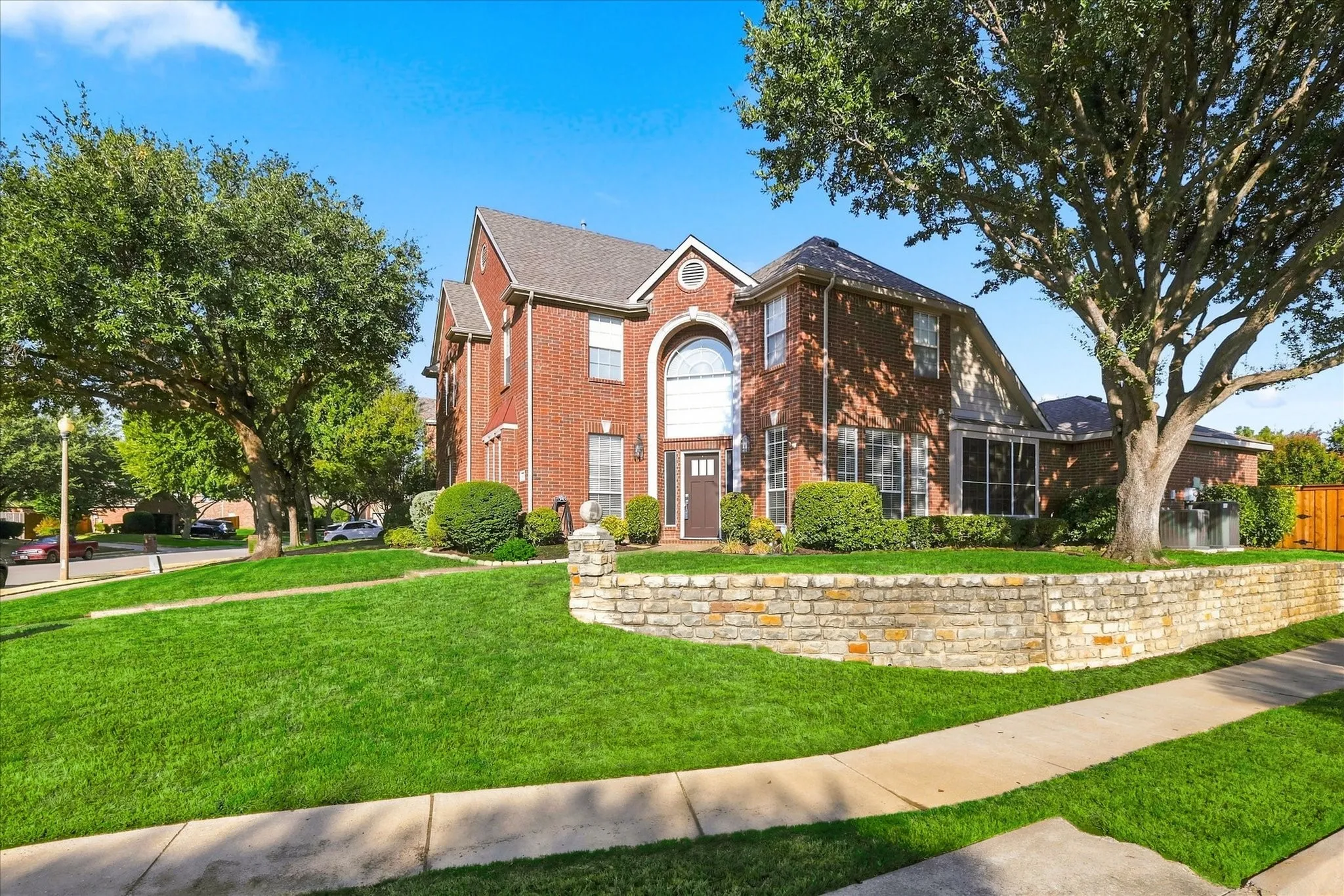 Brick exterior with a stone retaining wall, mature trees, and a well-maintained lawn