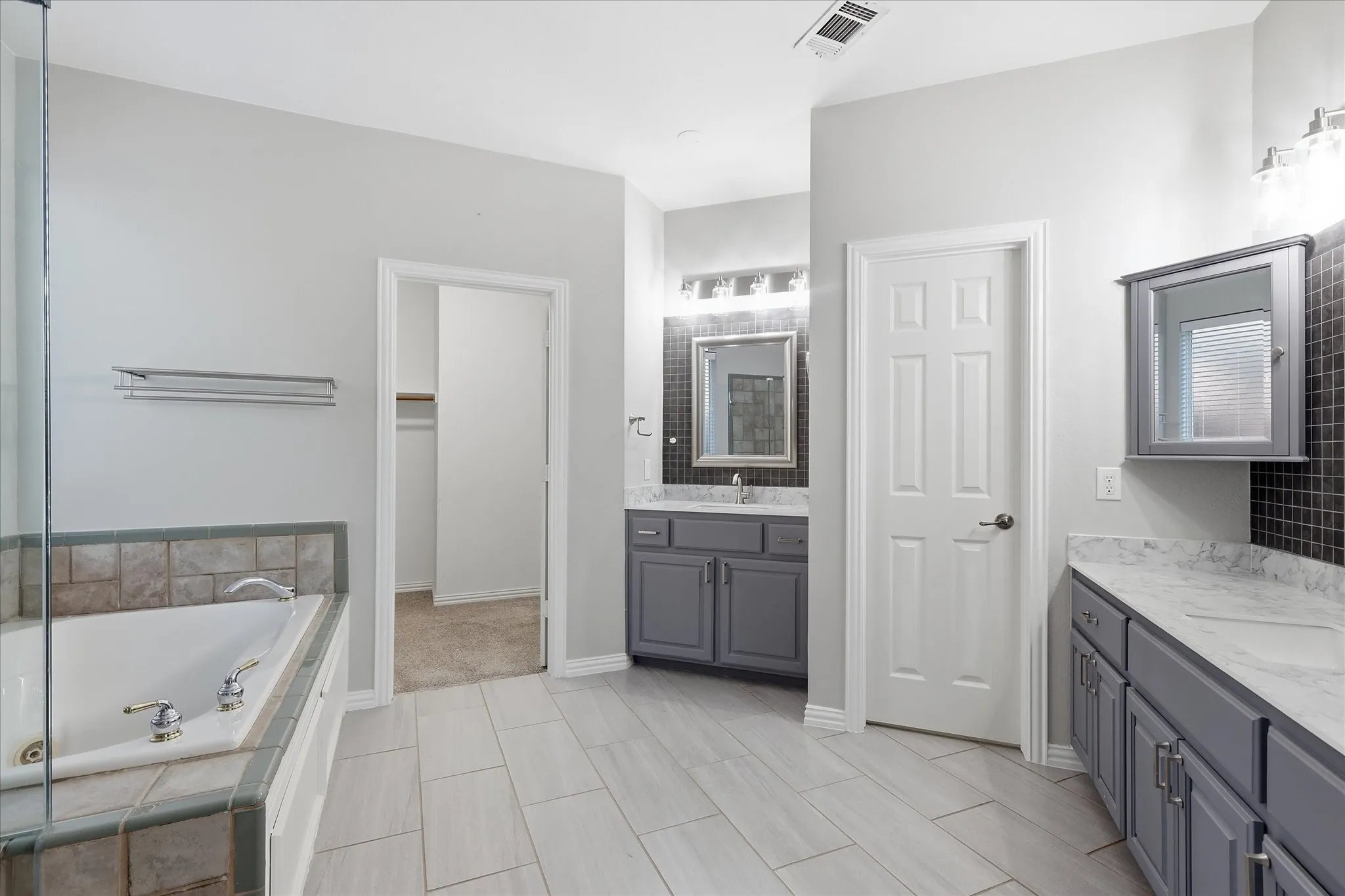 Dual vanity with stone-look countertops, built-in soaking tub with tile surround, glass-enclosed shower, light-toned plank-style flooring, and gray cabinetry
