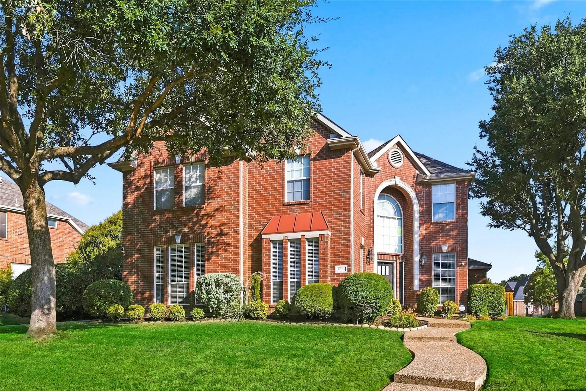 Brick exterior with a prominent arched window, mature trees, a manicured lawn, and a winding stone pathway