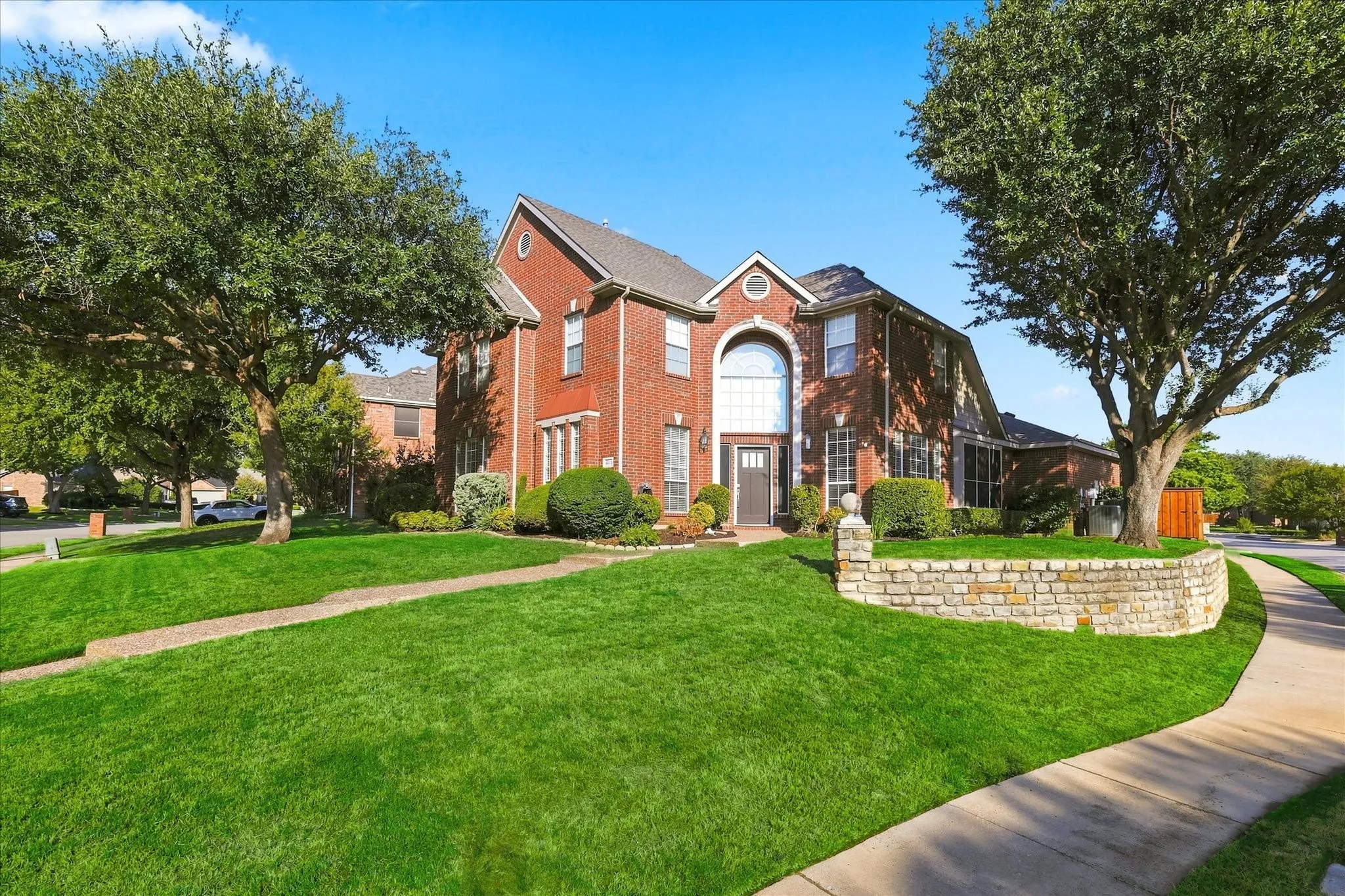 Brick exterior with a gabled roofline, arched window, stone retaining wall, and mature trees