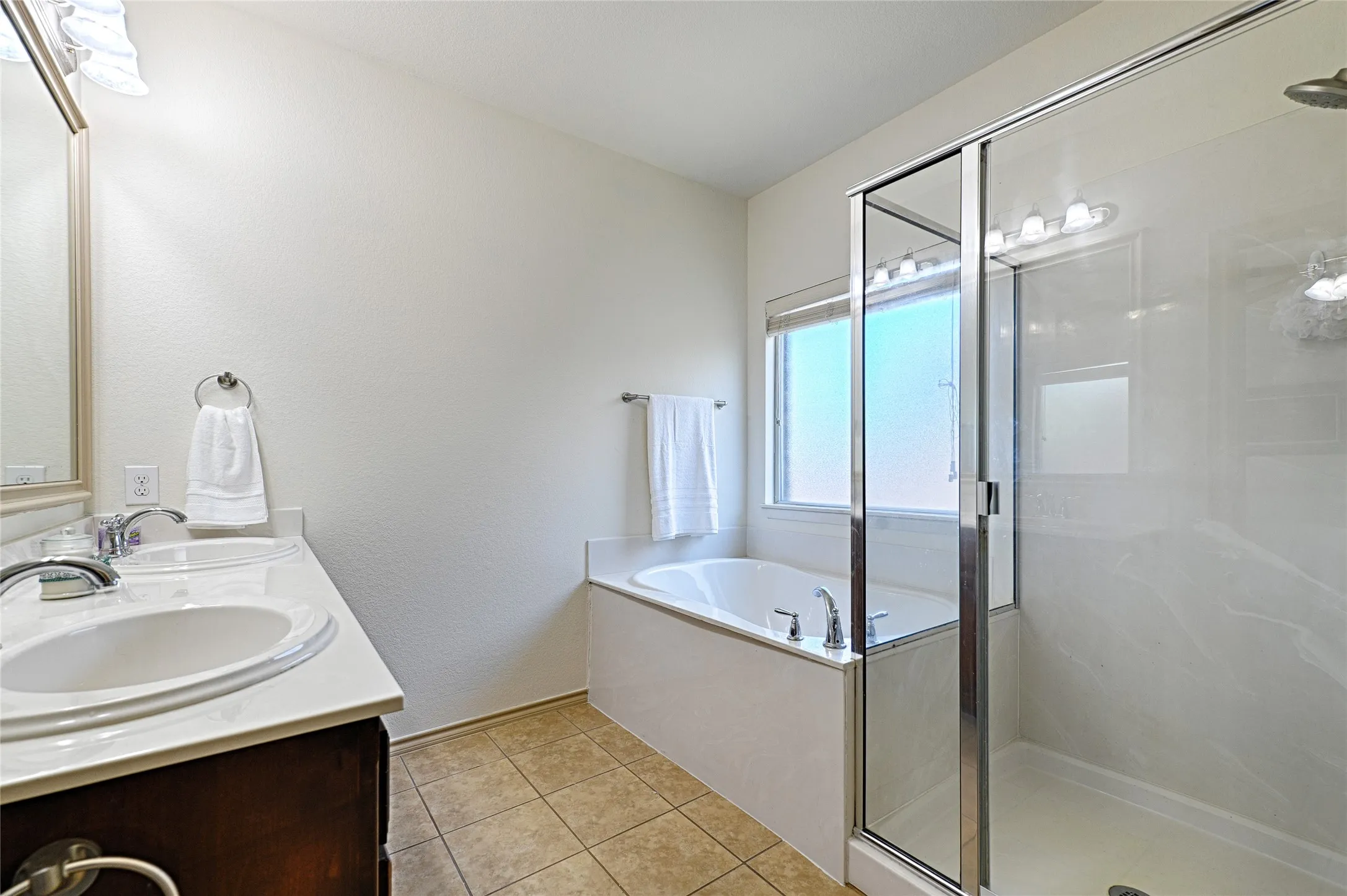 Bathroom featuring a spacious shower with a glass enclosure, a built-in bathtub with window, a double vanity with white countertops and undermount sinks, and tile flooring