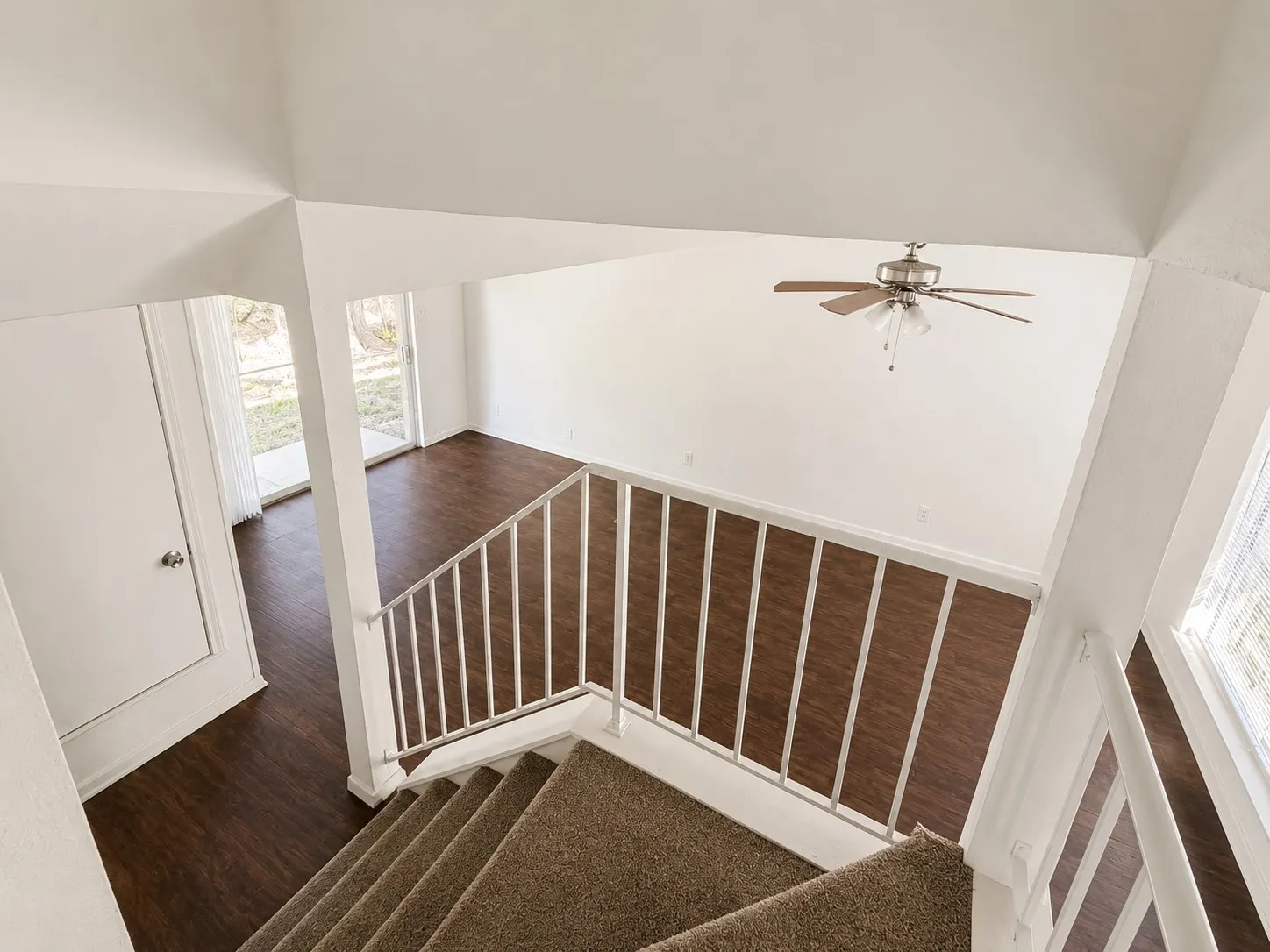 Carpeted staircase descending to a lower level featuring wood-finish flooring, white walls, a ceiling fan with light fixture, and a glass sliding door