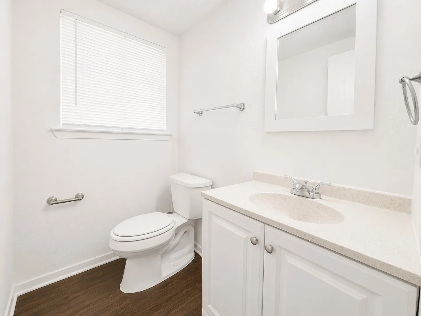 Half Guest Bathroom featuring a single window with blinds, a white vanity with an integrated sink and light countertop, chrome fixtures, dark wood-finish flooring, and a white toilet