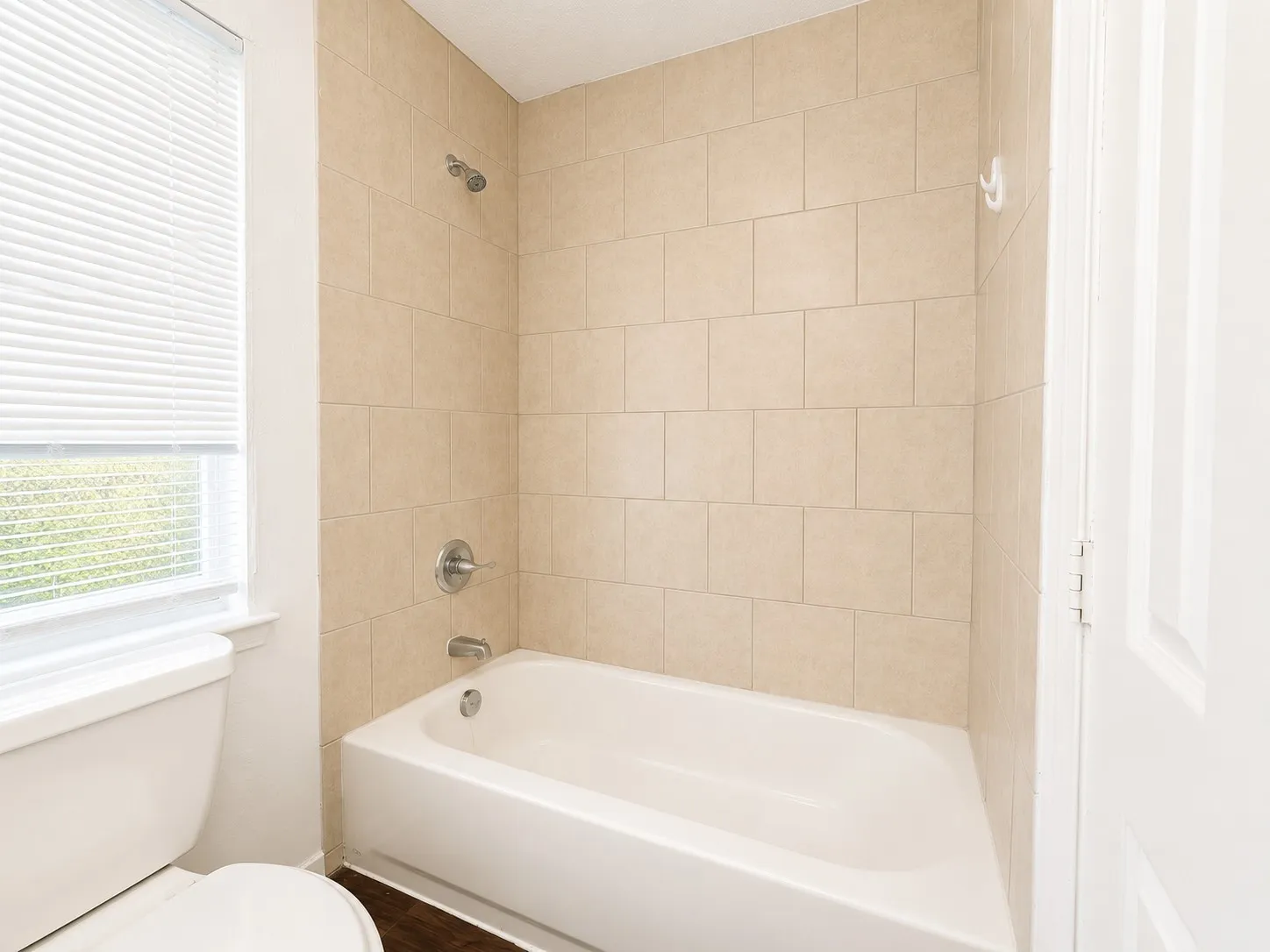 Bathroom featuring a bathtub with a shower head, tiled surround, white toilet, and a window with blinds