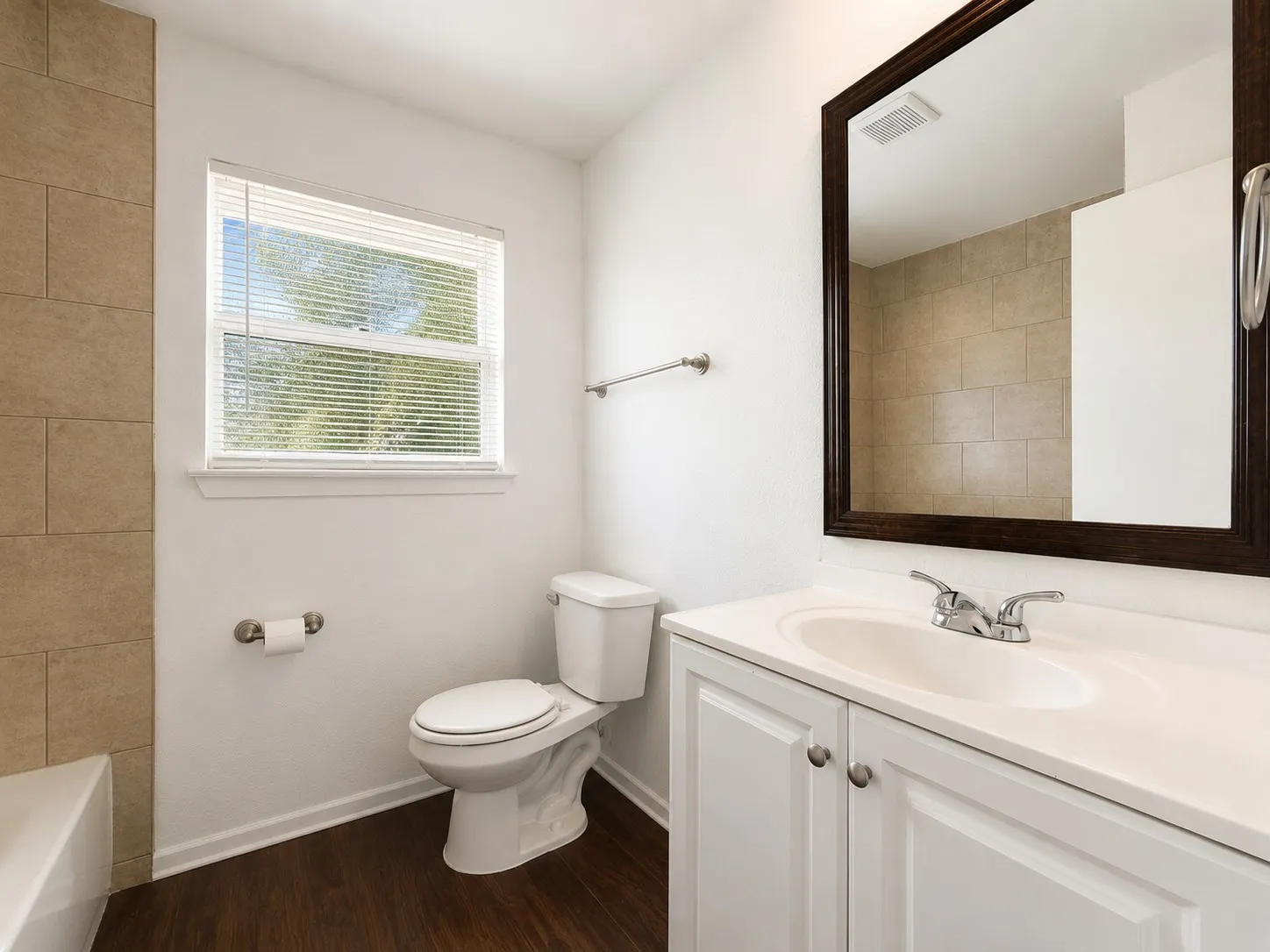 Bathroom featuring dark wood-finish flooring, a white vanity with integrated sink and chrome faucet, a framed mirror, a white toilet, and a window with blinds
