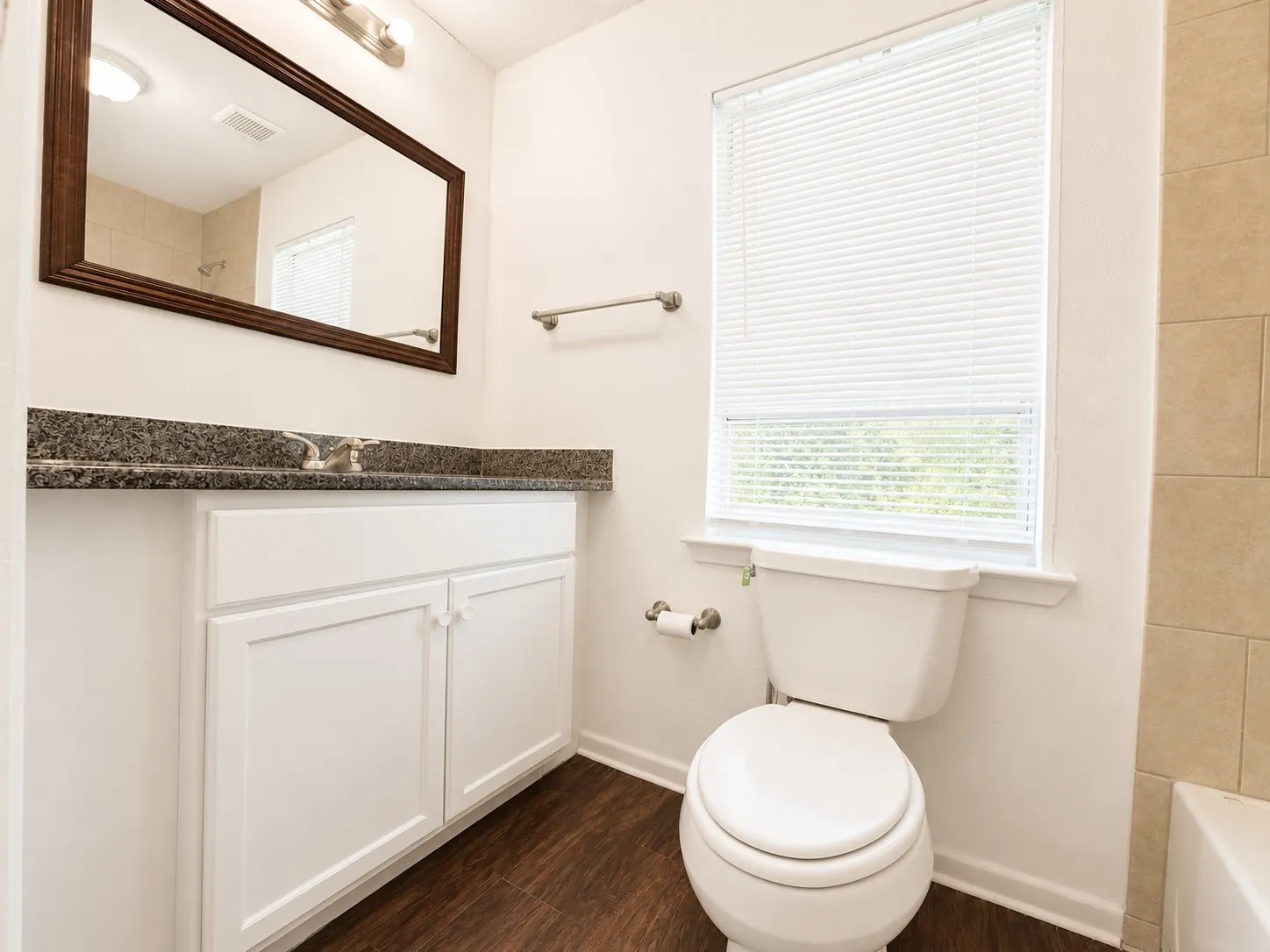 Bathroom featuring wood-finish flooring, a white vanity with paneled doors, a dark speckled granite countertop, and a window with blinds