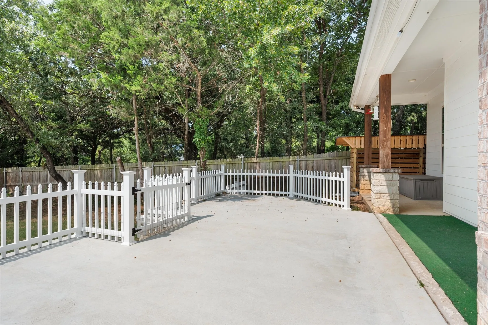Expansive concrete patio area featuring a white picket fence, a covered porch with stone columns and wood support beams, and mature trees providing natural screening