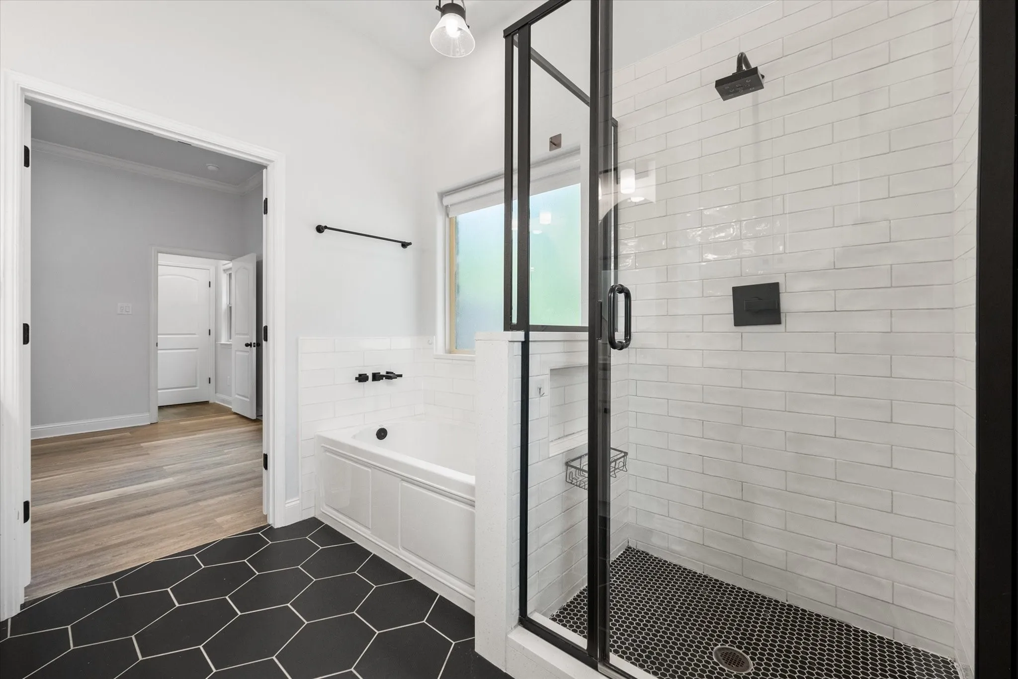 Black hexagon tile flooring with a white inset bathtub and a glass-enclosed shower featuring subway tile walls and a black mosaic tile base accent the primary bathroom