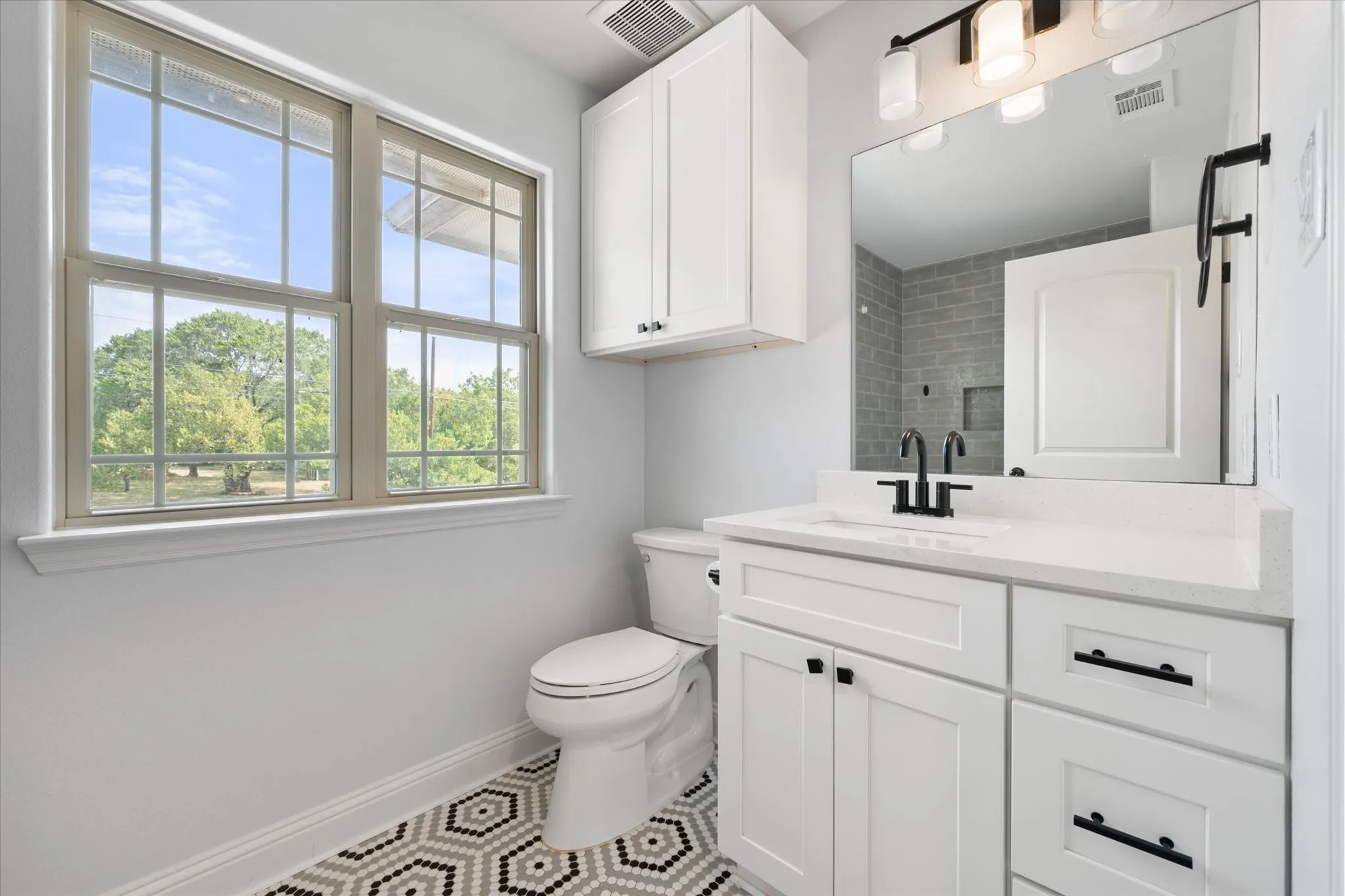 Windowed bathroom featuring a single vanity with quartz countertop, white shaker cabinetry with black hardware, attractive subway tiled tub shower and fun mosaic tiled floor