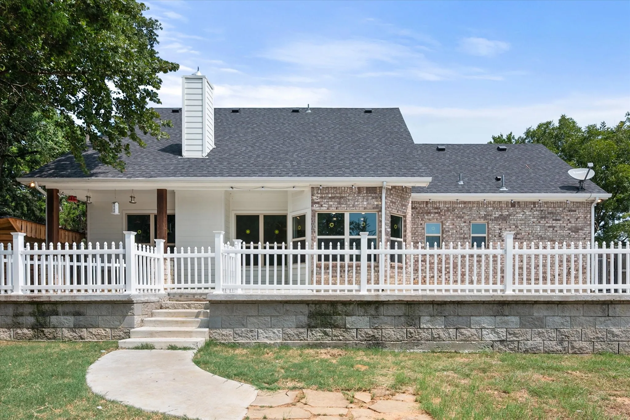Rear exterior featuring a covered patio, a white picket fence, a stone retaining wall, and a curved concrete pathway
