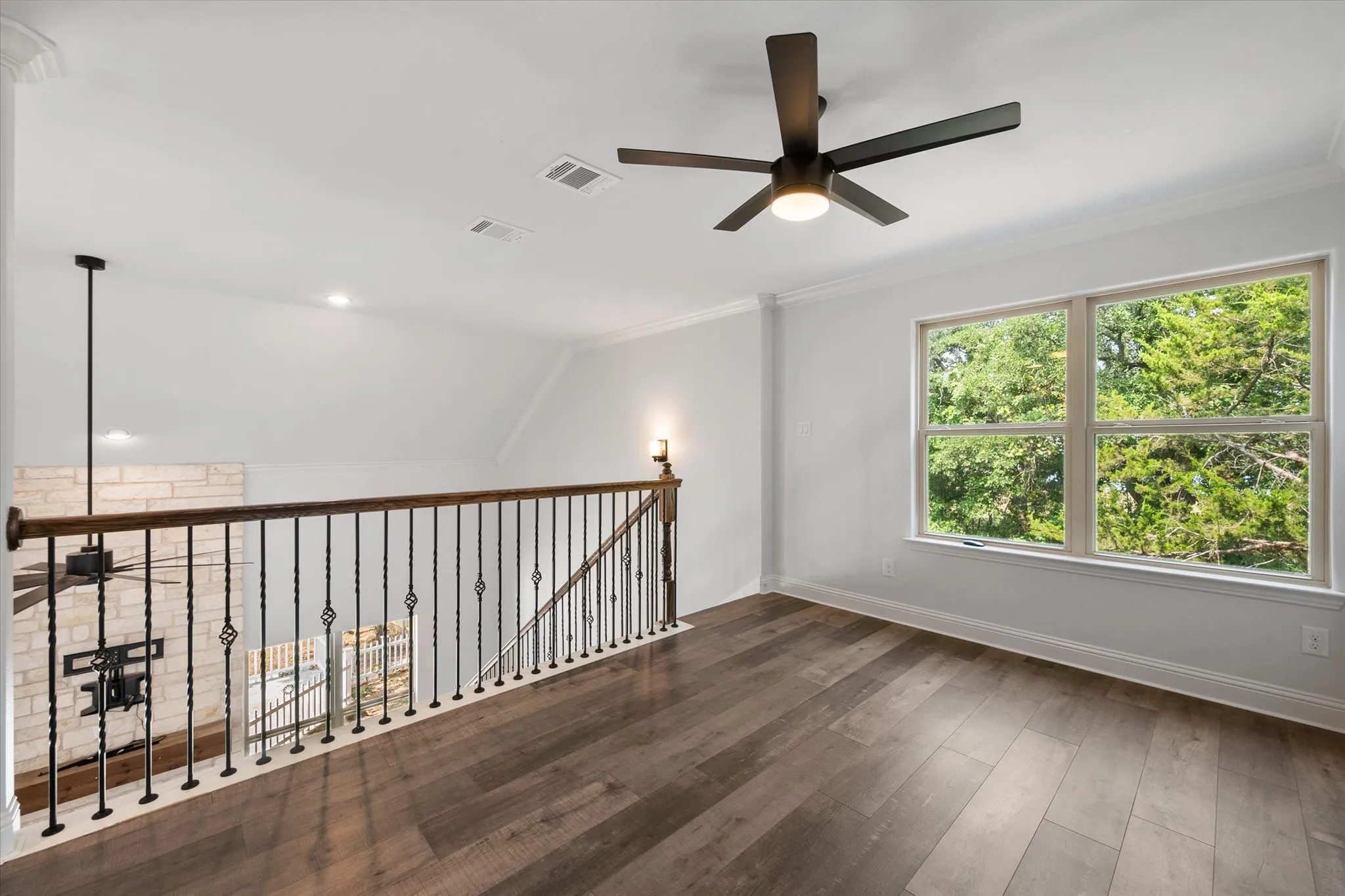 Loft area featuring wood-finish flooring, a contemporary ceiling fan, a large window with exterior foliage and views of the living area downstairs.