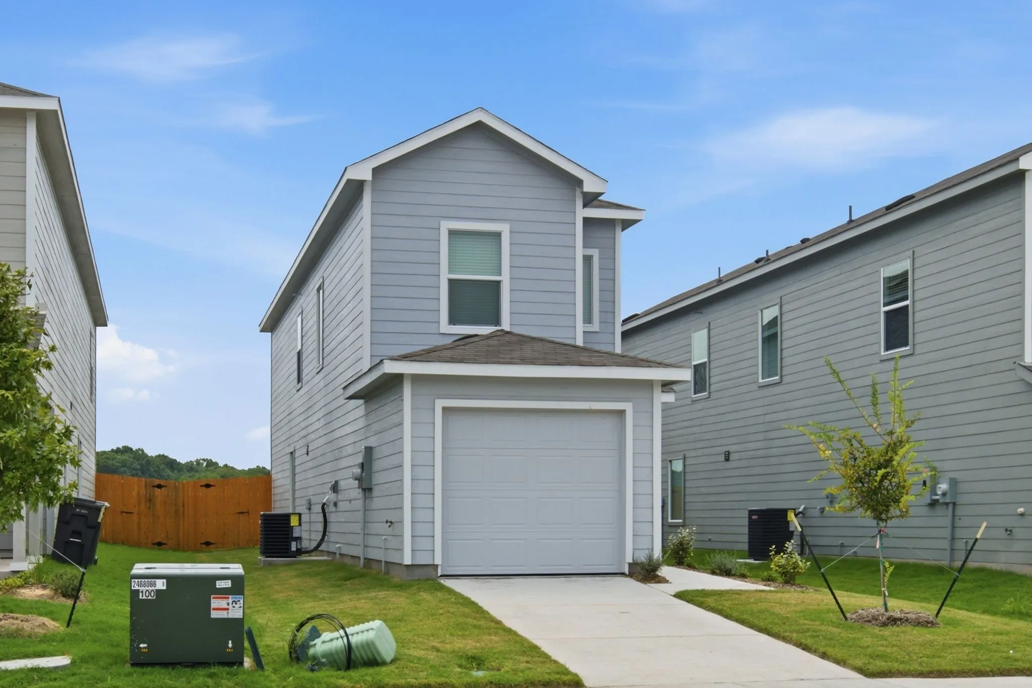 Two-story residence featuring light gray siding, an attached garage, a paved driveway, and a front lawn with a newly planted tree